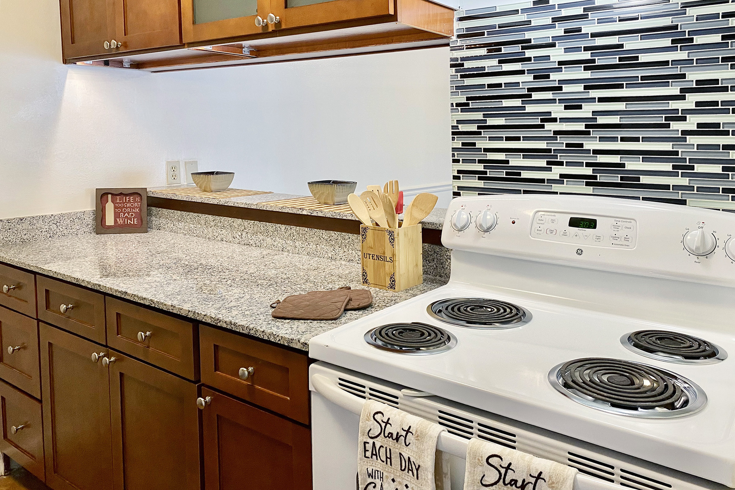 A modern kitchen featuring wooden cabinets, a granite countertop, and a glass-tiled backsplash. An electric stove is visible with cooking utensils and a decorative sign reading “Start each day with a smile.” The setting is bright and organized, ideal for cooking and meal preparation.