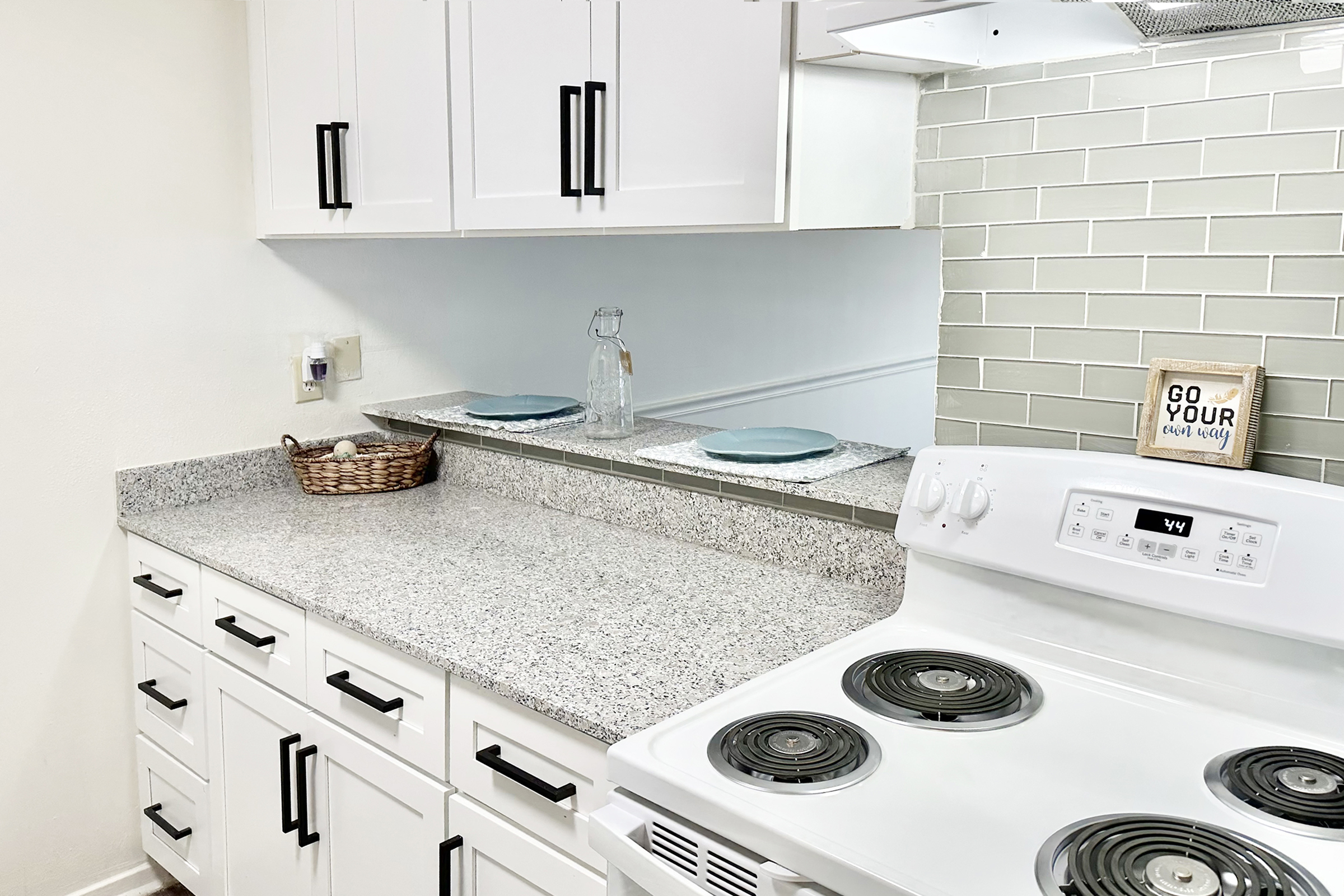 A bright kitchen featuring white cabinets with black handles, a granite countertop, and a stovetop. There is a glass water pitcher, two blue plates, and a woven basket with small items. A decorative sign reads "GO YOUR OWN WAY," and a mirrored backsplash reflects the kitchen's modern design.