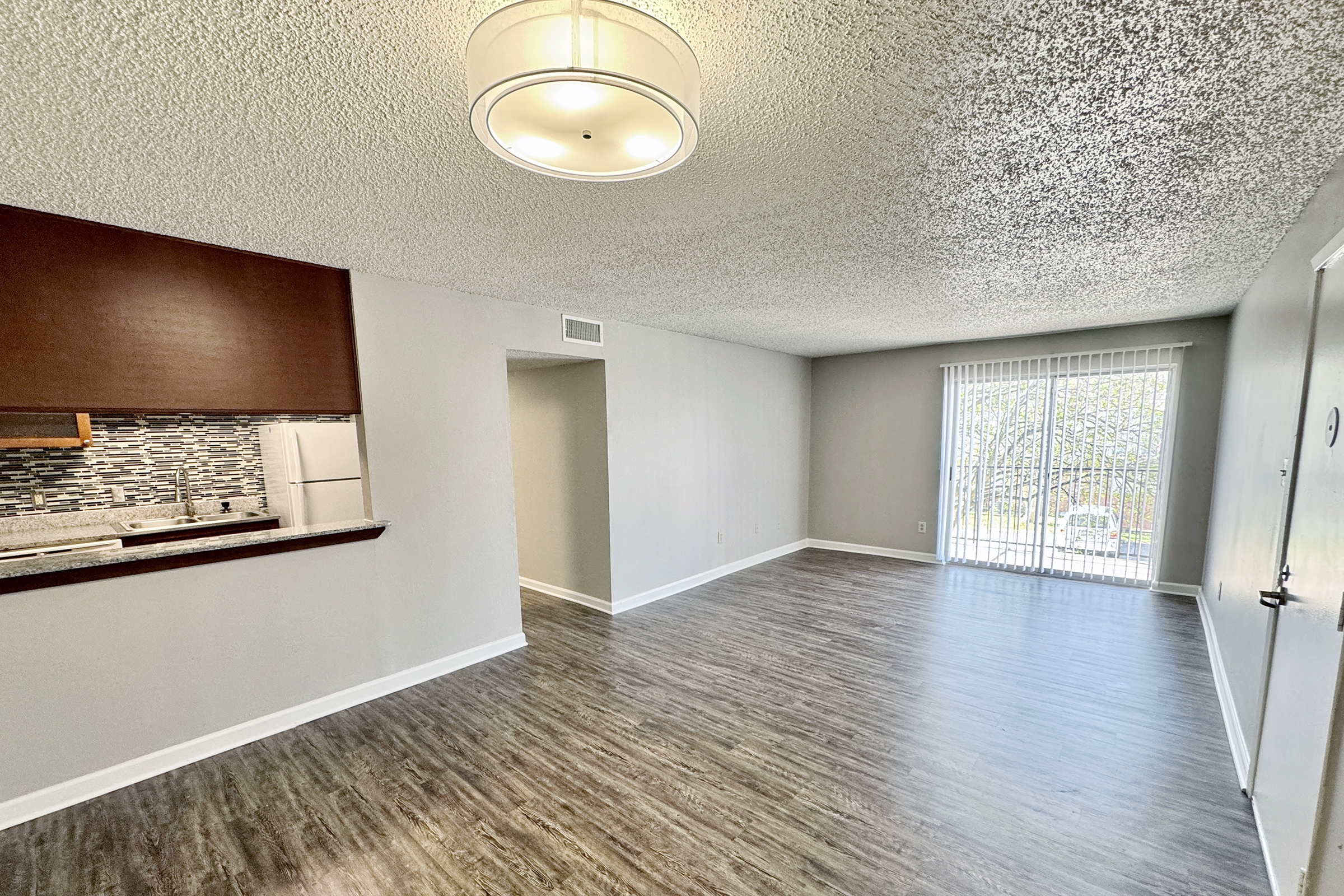 Spacious living area featuring light-colored walls, modern wood-style flooring, and a ceiling light fixture. To the left, a partial view of an open kitchen with a backsplash. Large sliding glass doors lead outside to a balcony or patio area, allowing natural light to fill the space.