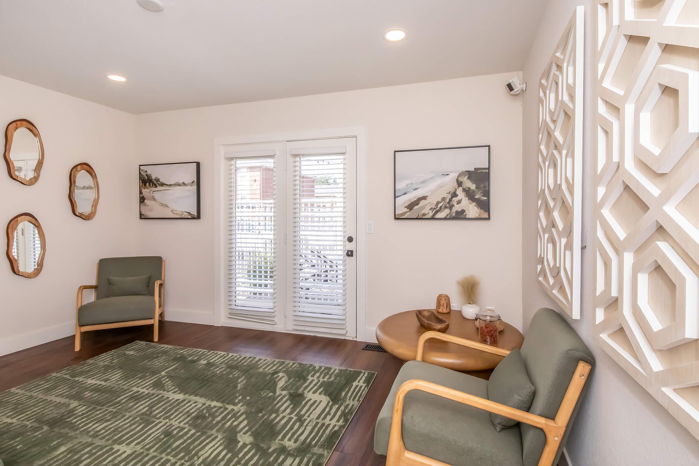 A cozy living room featuring two green armchairs, a round wooden coffee table, and decorative wall art including mirrors and framed nature prints. There are large windows with blinds allowing natural light to enter, and a textured green rug on the wooden floor.