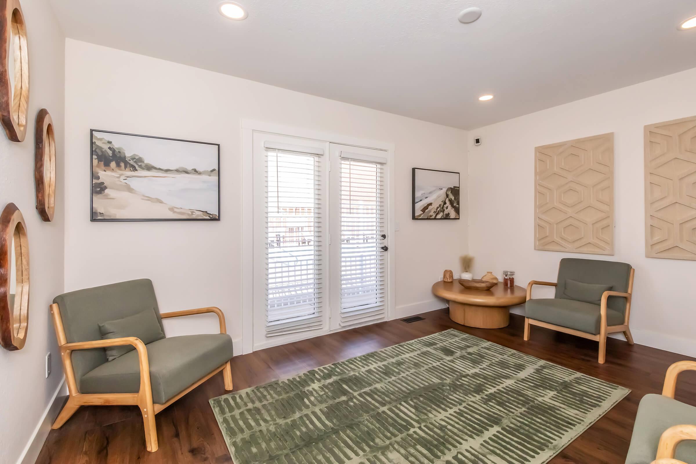 A cozy living room featuring two green armchairs, a round wooden coffee table, and a textured area rug. The walls are adorned with two framed art pieces, and there are large sliding glass doors with blinds that lead to an outdoor space, creating a bright and inviting atmosphere.