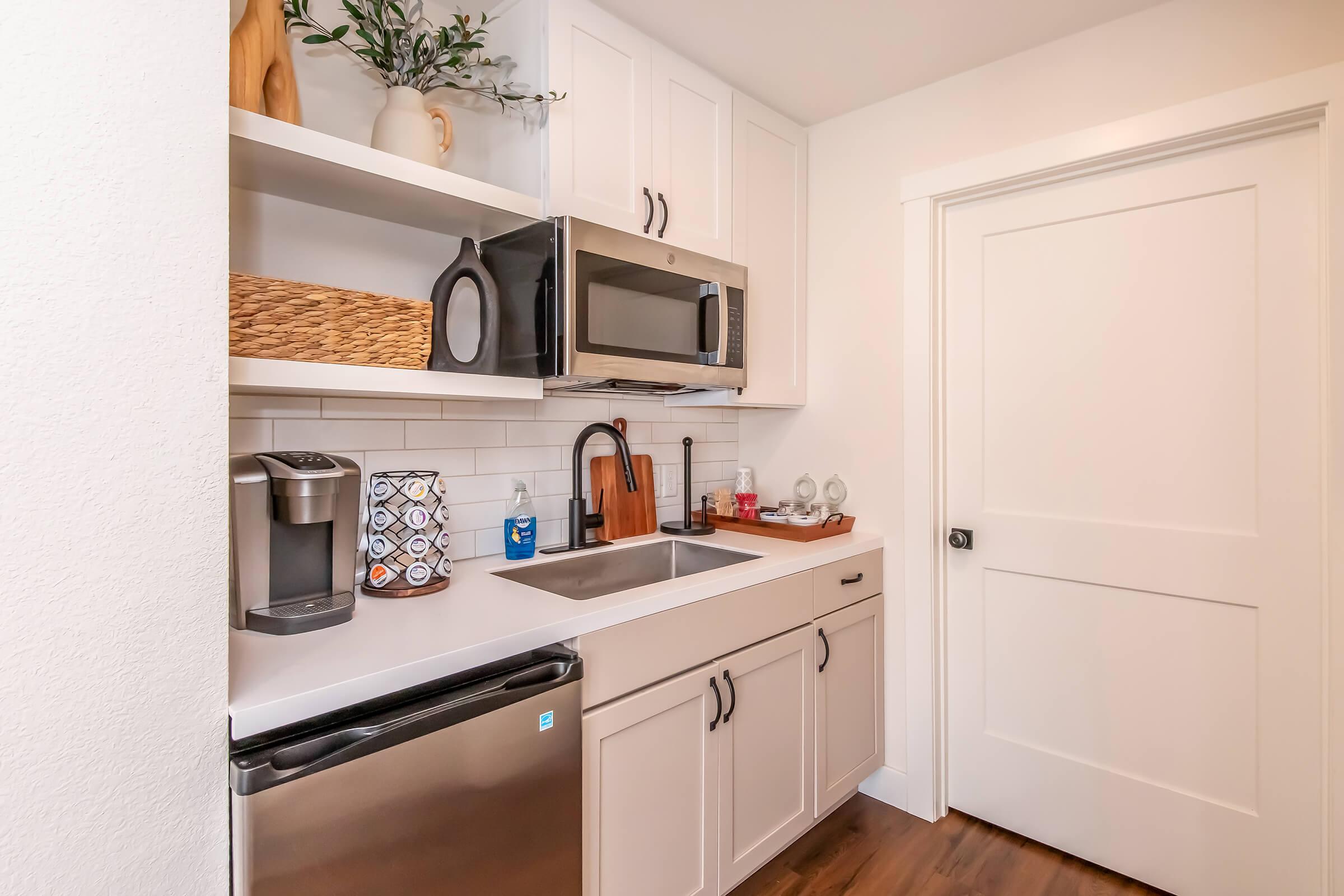 A modern kitchen featuring white cabinetry, a sink, a microwave, and a coffee maker. There are decorative items on the shelves, including a plant and a woven basket. A stainless steel refrigerator is visible, along with a closed door in the background. The overall aesthetic is clean and stylish.