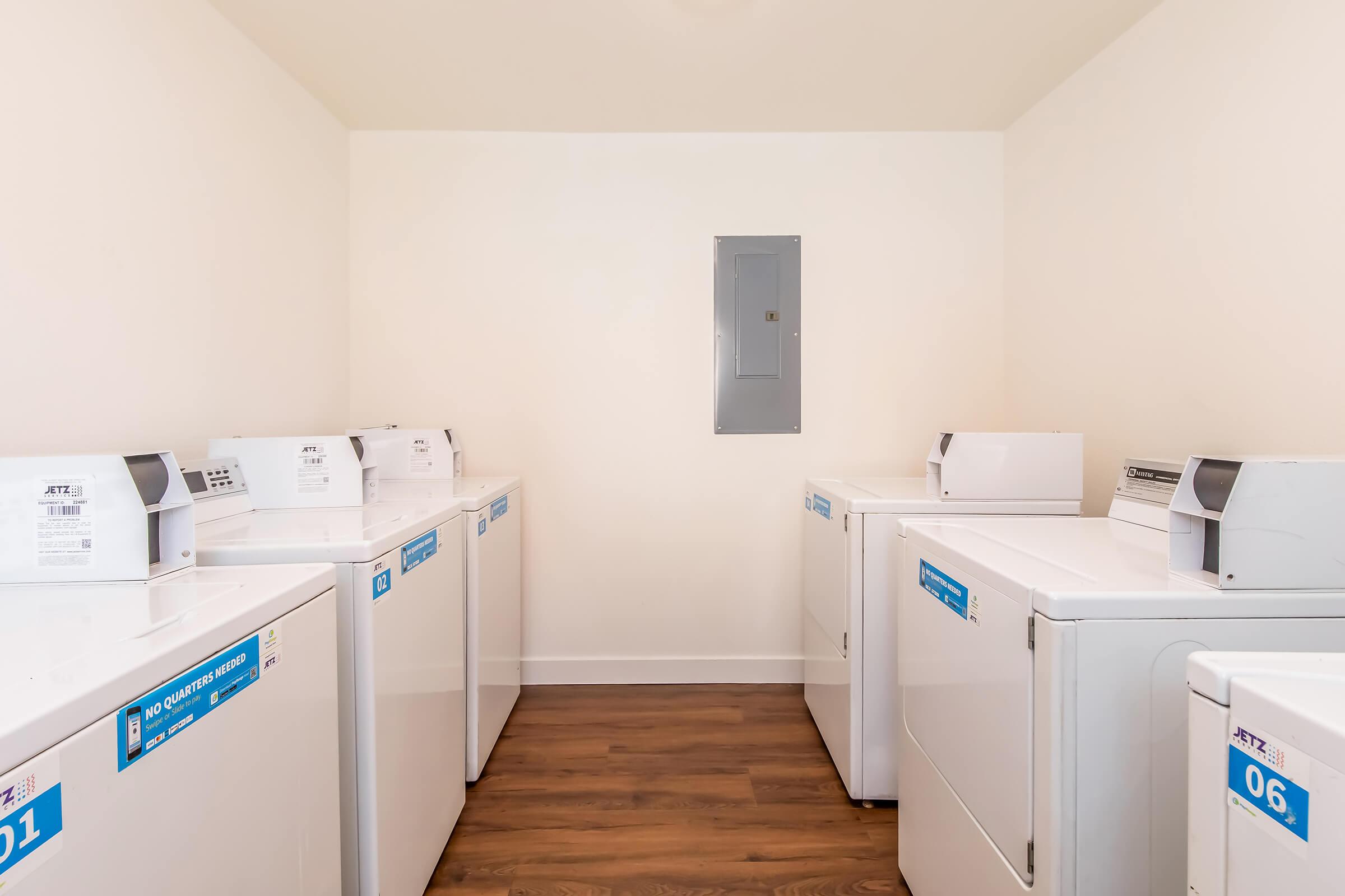 A clean and well-lit laundry room featuring several white washers and dryers lined up against the walls. Each machine has a numbered label. The floor is wooden, and there is a utility panel on the back wall. The overall space is tidy and organized.