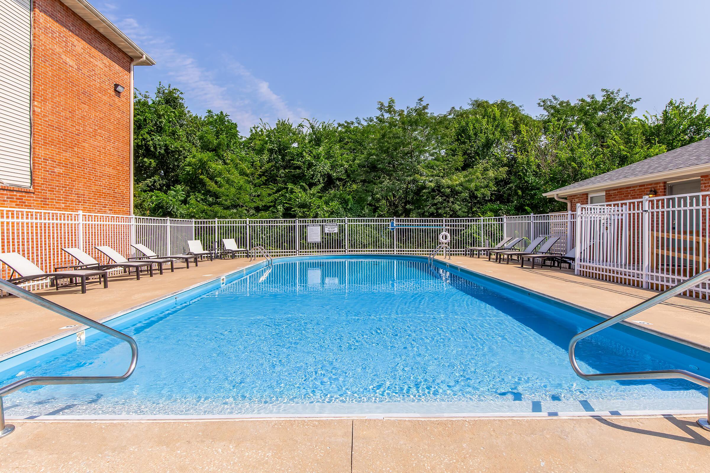A bright outdoor swimming pool surrounded by a metal fence. Lounge chairs are positioned around the pool on a concrete deck. In the background, lush green trees can be seen, and a small building is visible to the right. Clear blue sky overhead.