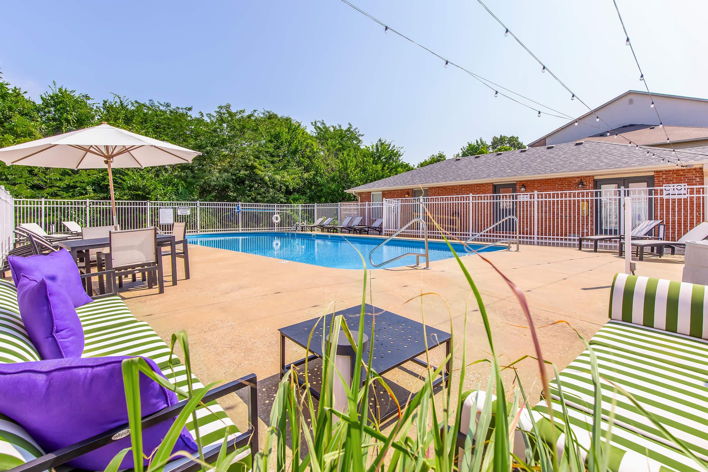 A sunny outdoor pool area featuring a clear blue pool surrounded by lounge chairs with green and white stripes. There is a large umbrella providing shade, and the space is bordered by greenery and a fenced area. String lights are hung overhead, enhancing the inviting atmosphere.