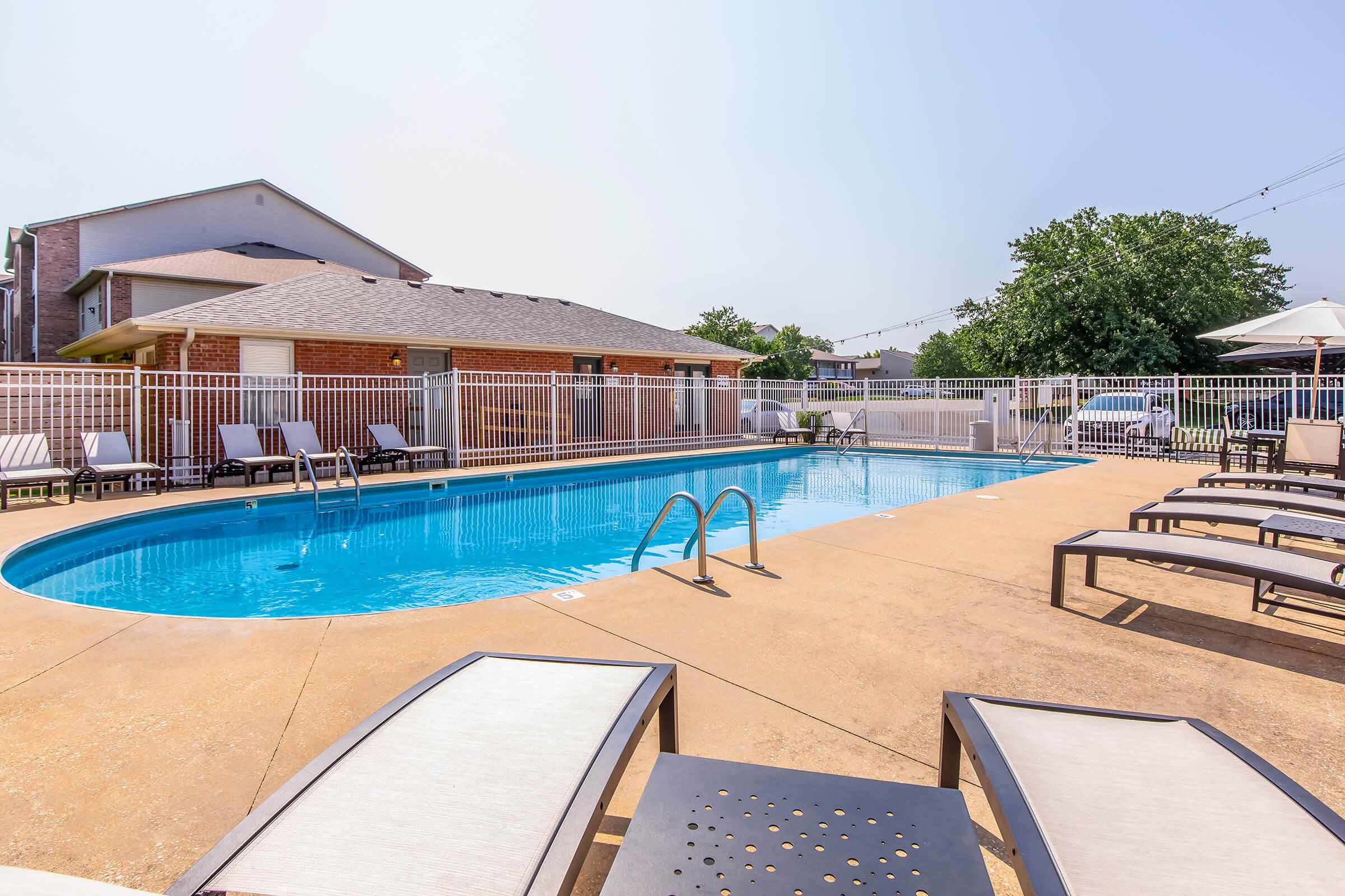 A clear blue swimming pool surrounded by lounge chairs and a fence, with a residential building in the background and trees nearby. The sky is bright and sunny, creating a warm, inviting atmosphere.
