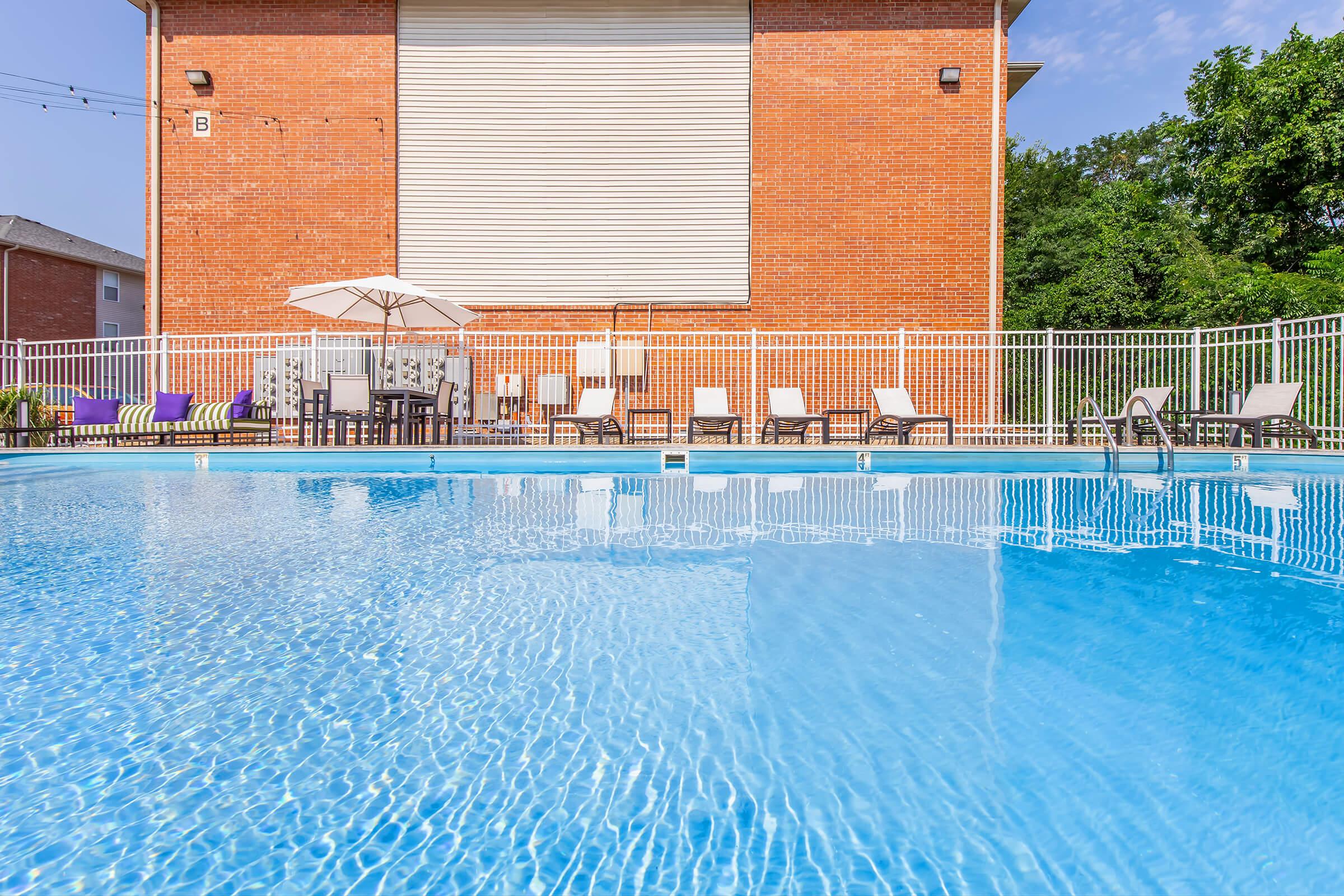 A swimming pool with clear blue water surrounded by lounge chairs and umbrellas. There is a brick building in the background with a roll-up door and green trees nearby. The scene is bright and inviting, ideal for relaxation and leisure activities.