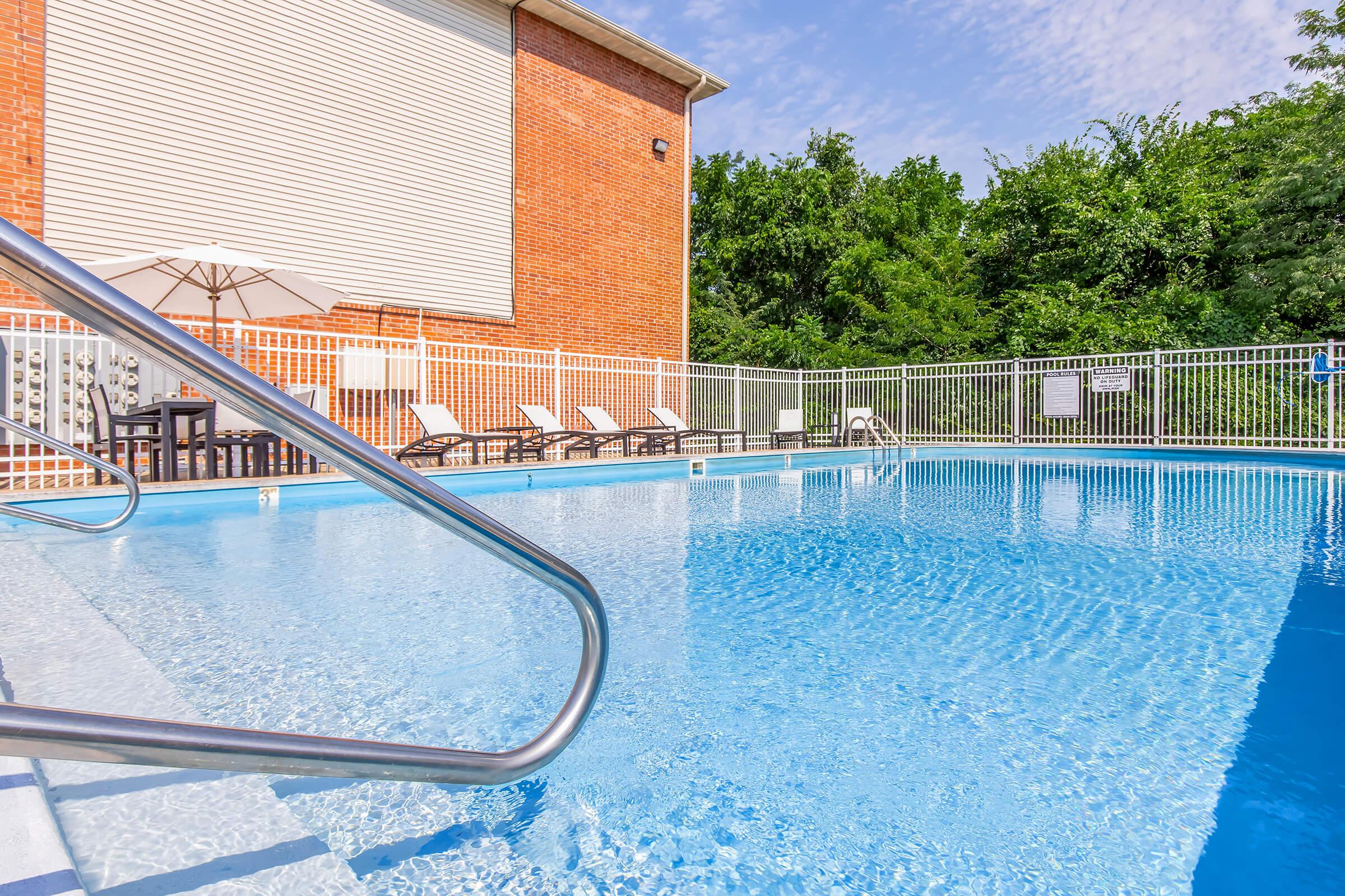 A clear blue swimming pool surrounded by a white fence, with several lounge chairs positioned nearby. An umbrella provides shade for seating. In the background, there are lush green trees and a brick building with a white exterior. The sky is bright and partly cloudy.