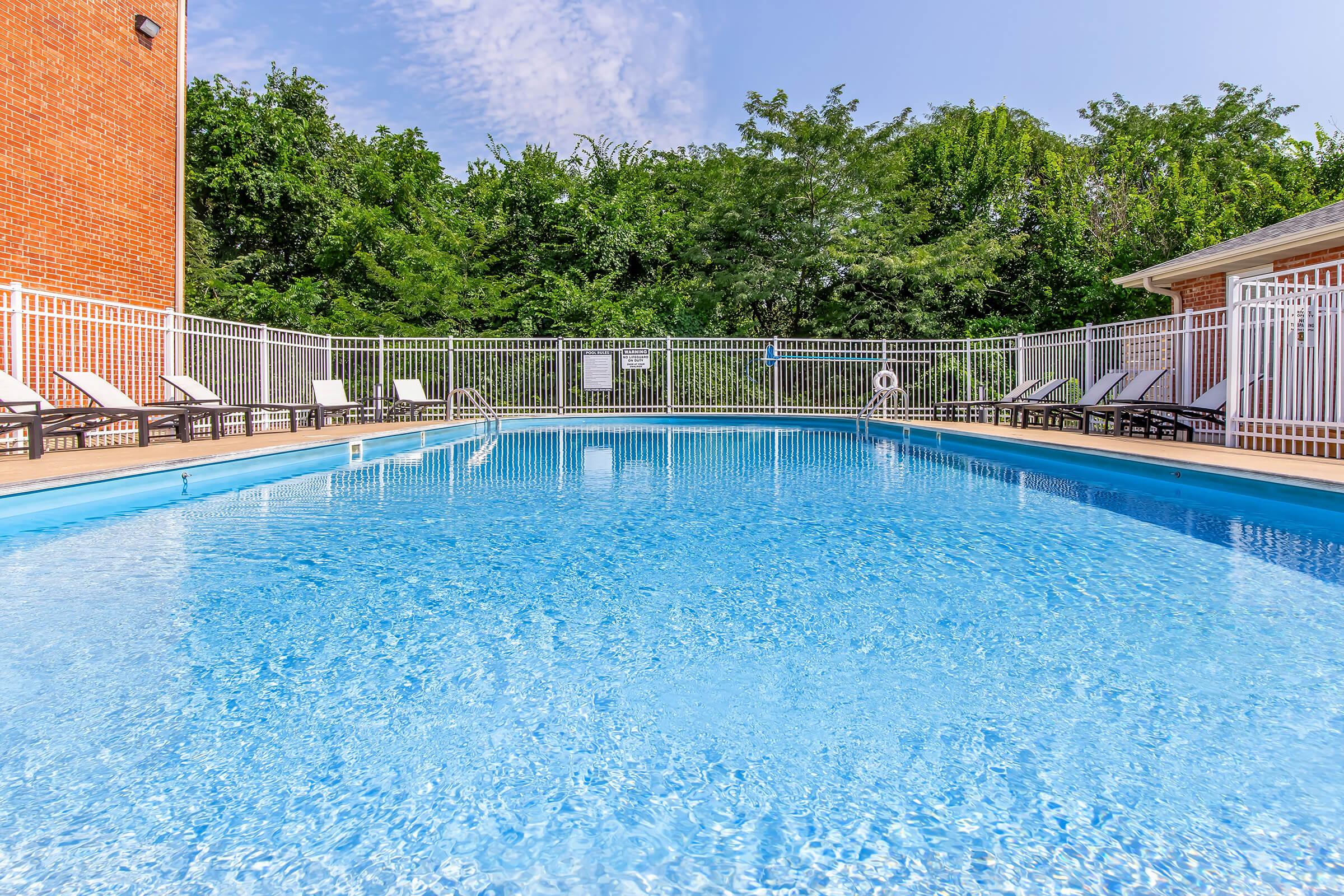 A clear blue swimming pool surrounded by lounge chairs and a white fence, with lush green trees in the background and a bright blue sky. The scene reflects a peaceful and inviting outdoor space for relaxation and leisure.