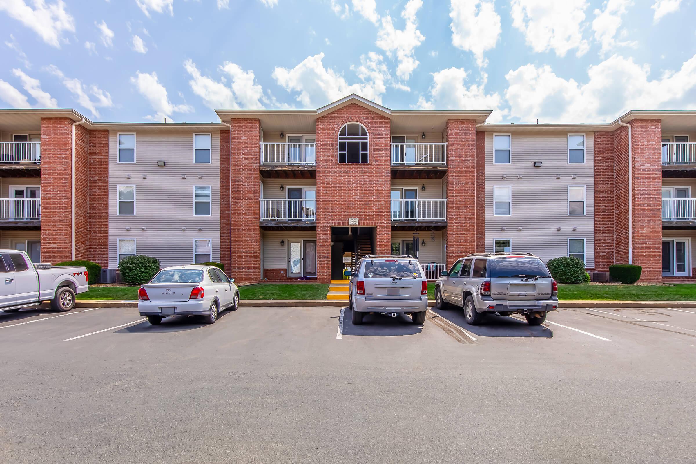 Apartment building featuring three stories with a brick facade, balconies, and multiple vehicles parked in front. The sky is partly cloudy, and the setting includes a well-maintained parking area.