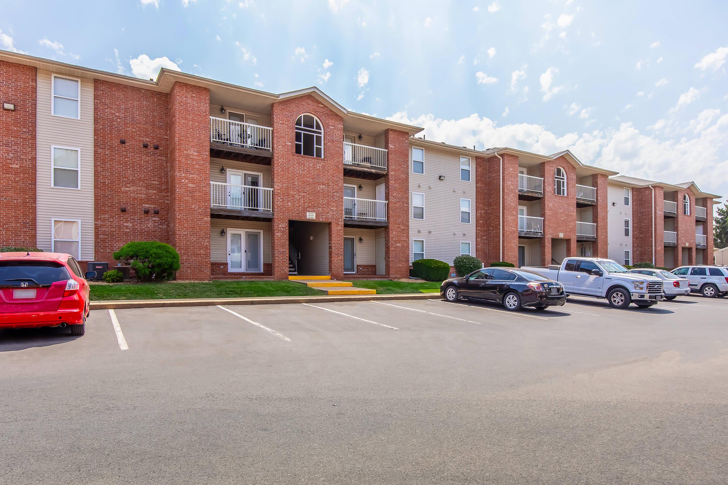A view of a multi-unit residential apartment building with brick exterior. The building features balconies and a well-maintained lawn. Several parking spaces are visible in front, with a red car and a few other vehicles parked. The sky is partly cloudy, indicating a sunny day.