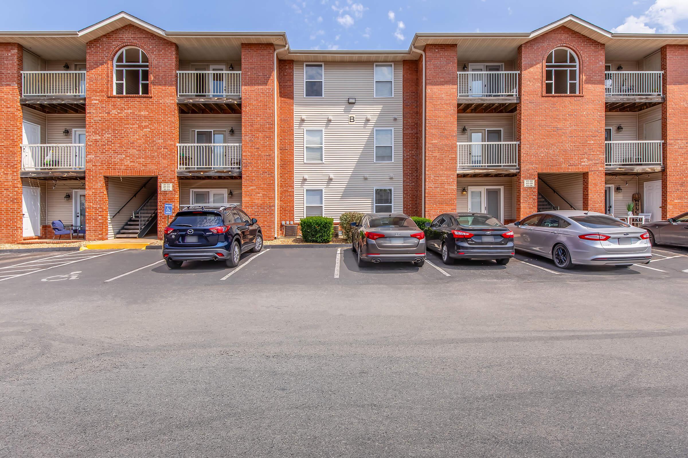 A brick apartment building with three floors and balconies, featuring several parked cars in front. The parking lot is lined with marked spaces, and the building has a clean, well-maintained appearance under a clear blue sky.