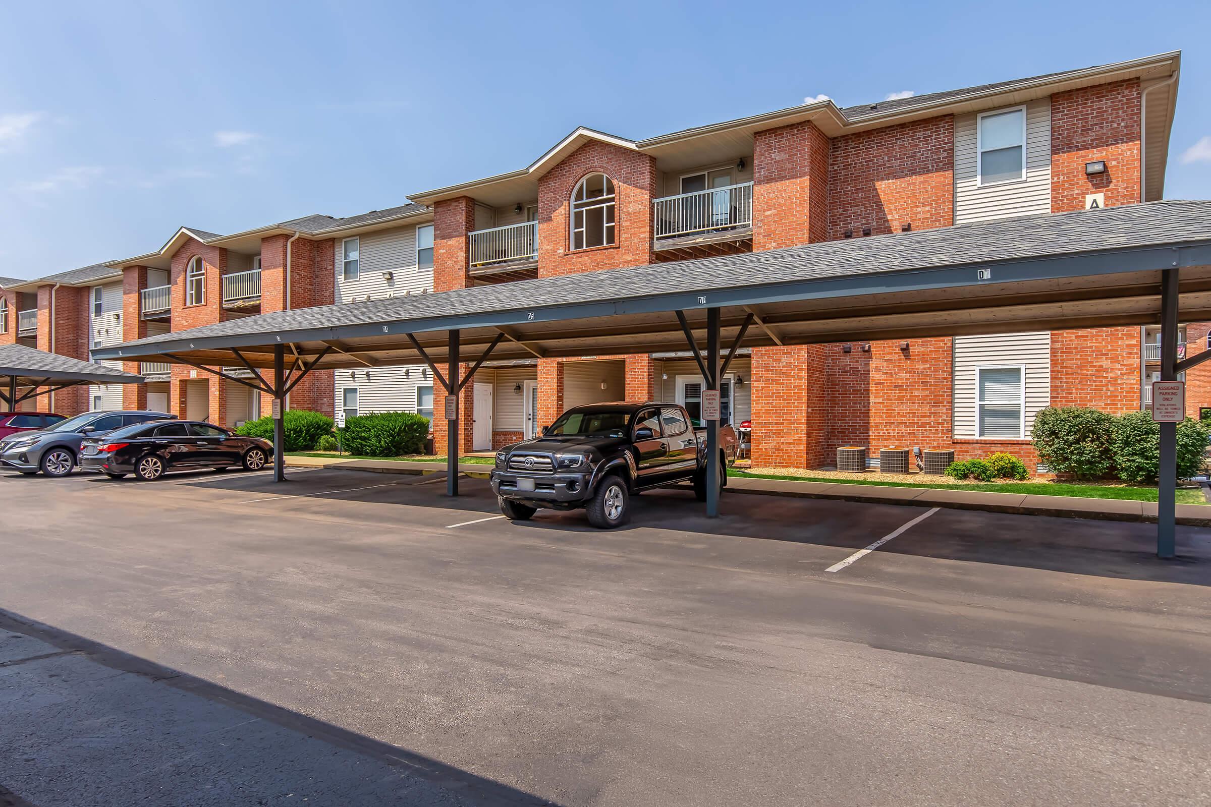 Apartment complex featuring multiple units with brick exteriors and covered parking spaces. Several cars are parked under the awnings, with well-maintained landscaping and a clear blue sky in the background. The scene conveys a tidy and inviting residential environment.