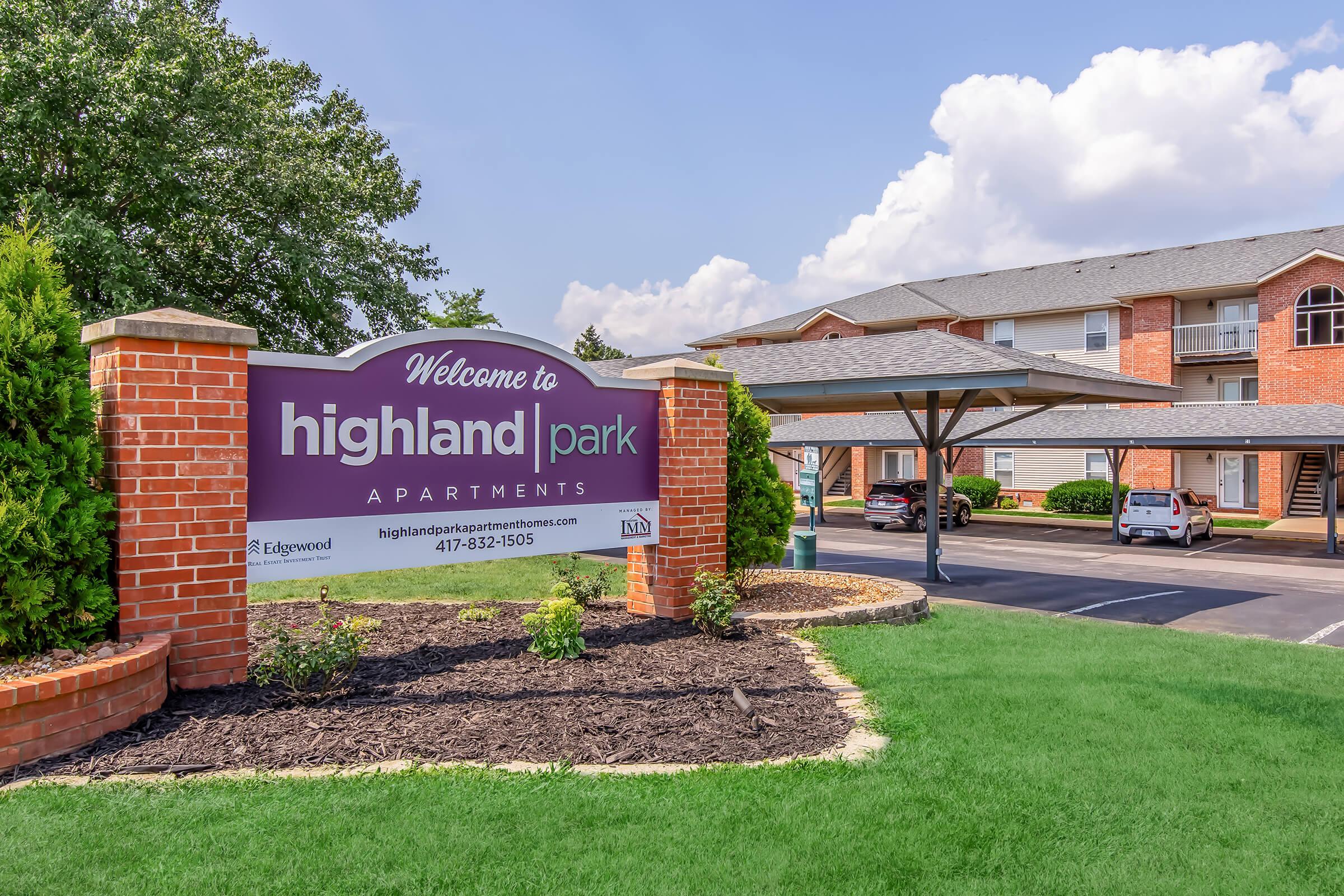 Welcome sign for Highland Park Apartments, featuring brick pillars and landscaped greenery. In the background, apartment buildings are visible, along with a car parked under a canopy. The sky is partly cloudy, creating a bright atmosphere.
