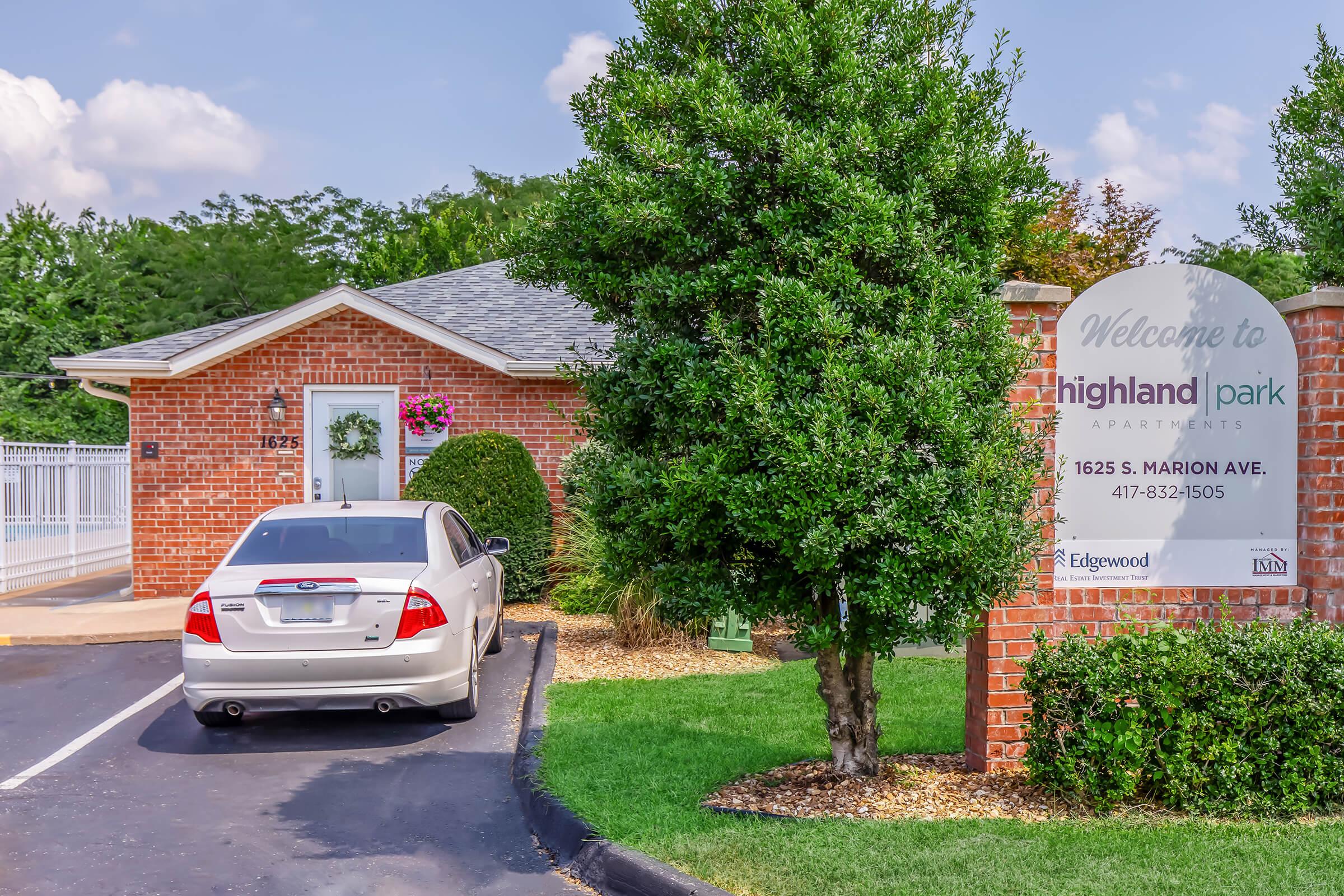 A brick building with a sign that reads "Welcome to Highland Park Apartments" next to a parked silver car. The entrance features a decorative wreath and well-maintained landscaping including bushes and grass, under a partly cloudy sky. A clear view of the apartment's address is visible.