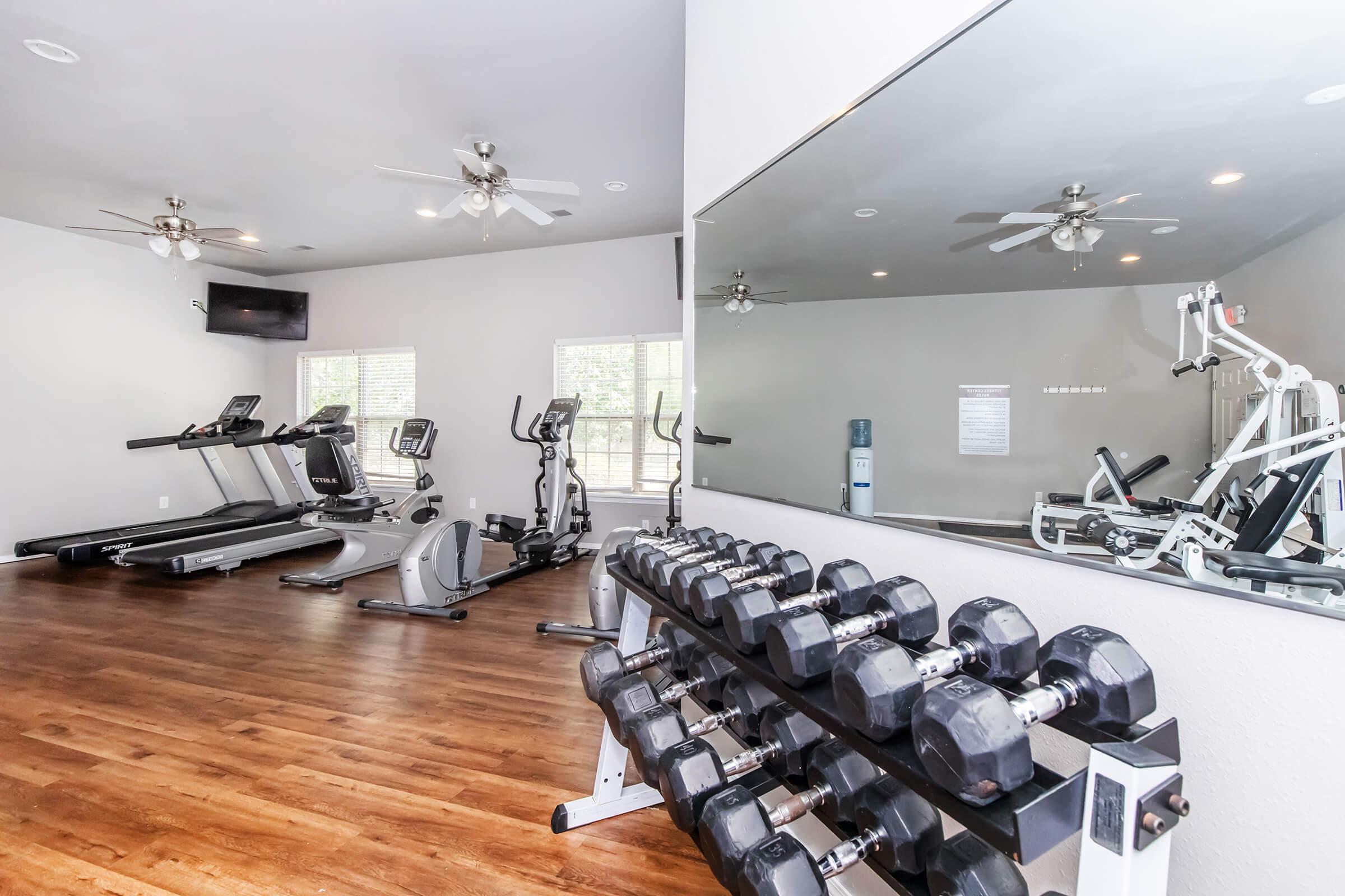 A modern gym featuring a variety of fitness equipment. In the foreground, a rack of dumbbells is visible. In the background, there are treadmills, an elliptical machine, and a multi-station weight machine. Large windows provide natural light, and the flooring is made of hardwood.
