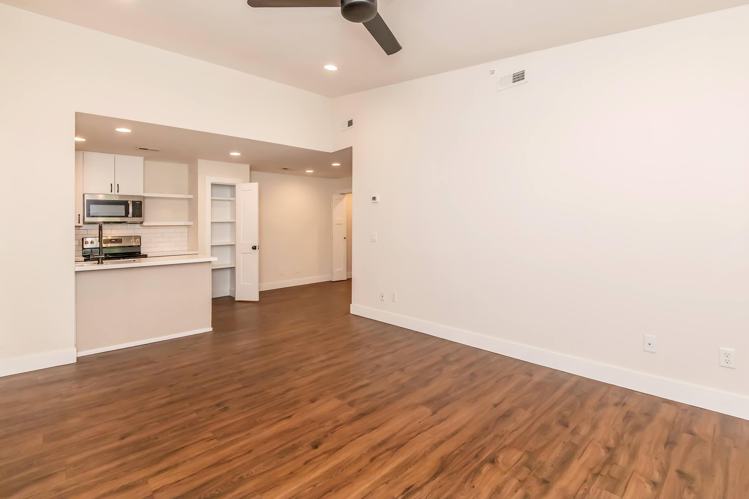 Bright, empty living space featuring a modern design with light-colored walls, hardwood flooring, a ceiling fan, and an open kitchen area in the background. There are built-in shelves and a doorway leading to another room. Overall, the space is clean and ready for furniture.