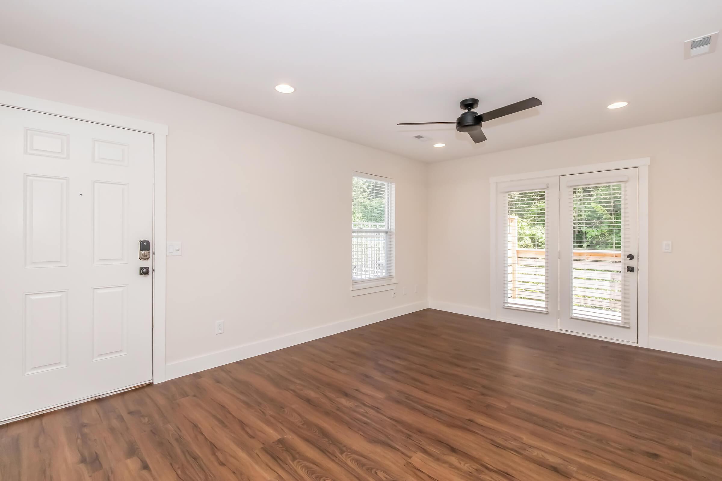 Bright and spacious living room with a ceiling fan, wood flooring, and white walls. It features a front door and sliding glass doors that open to a balcony or patio, allowing natural light to fill the space. Modern design with no furniture, creating an open and inviting atmosphere.