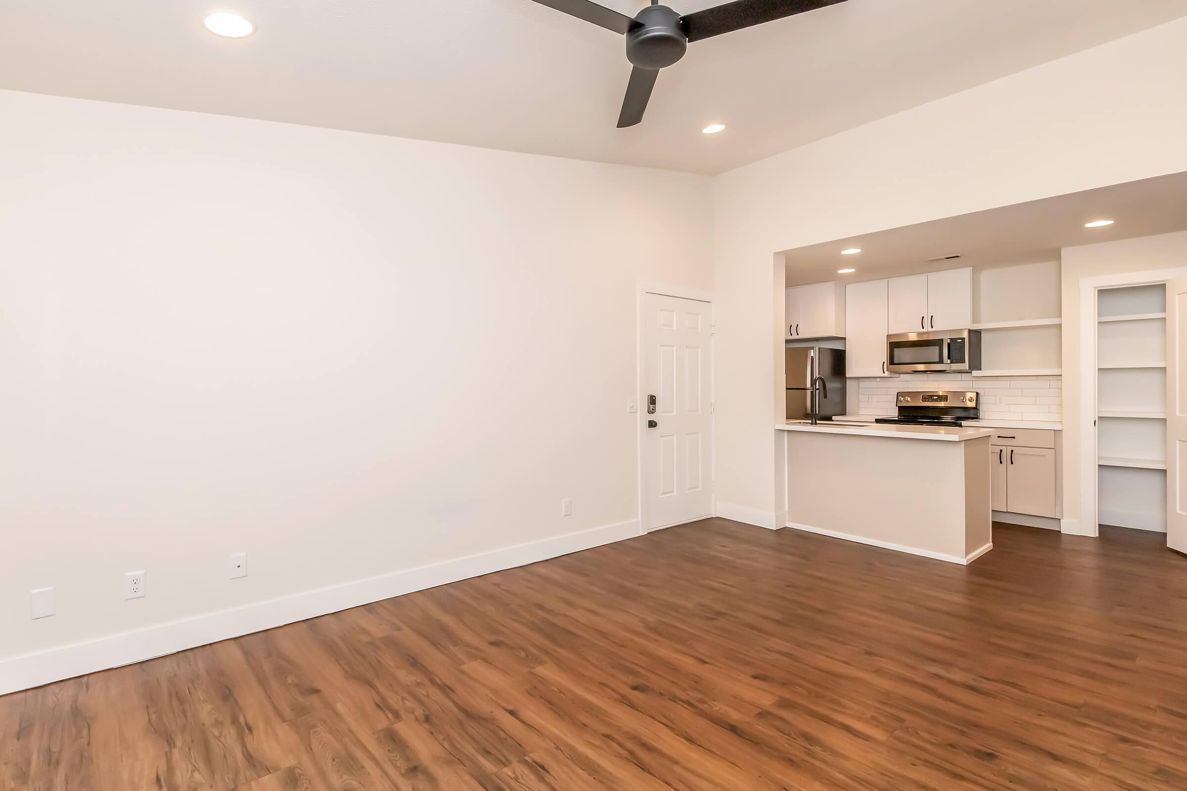 A spacious, modern living area featuring light-colored walls, a ceiling fan, and hardwood flooring. To the right, there is an open kitchen with stainless steel appliances and white cabinetry. The entry door is visible, and the space is well-lit with recessed lighting.