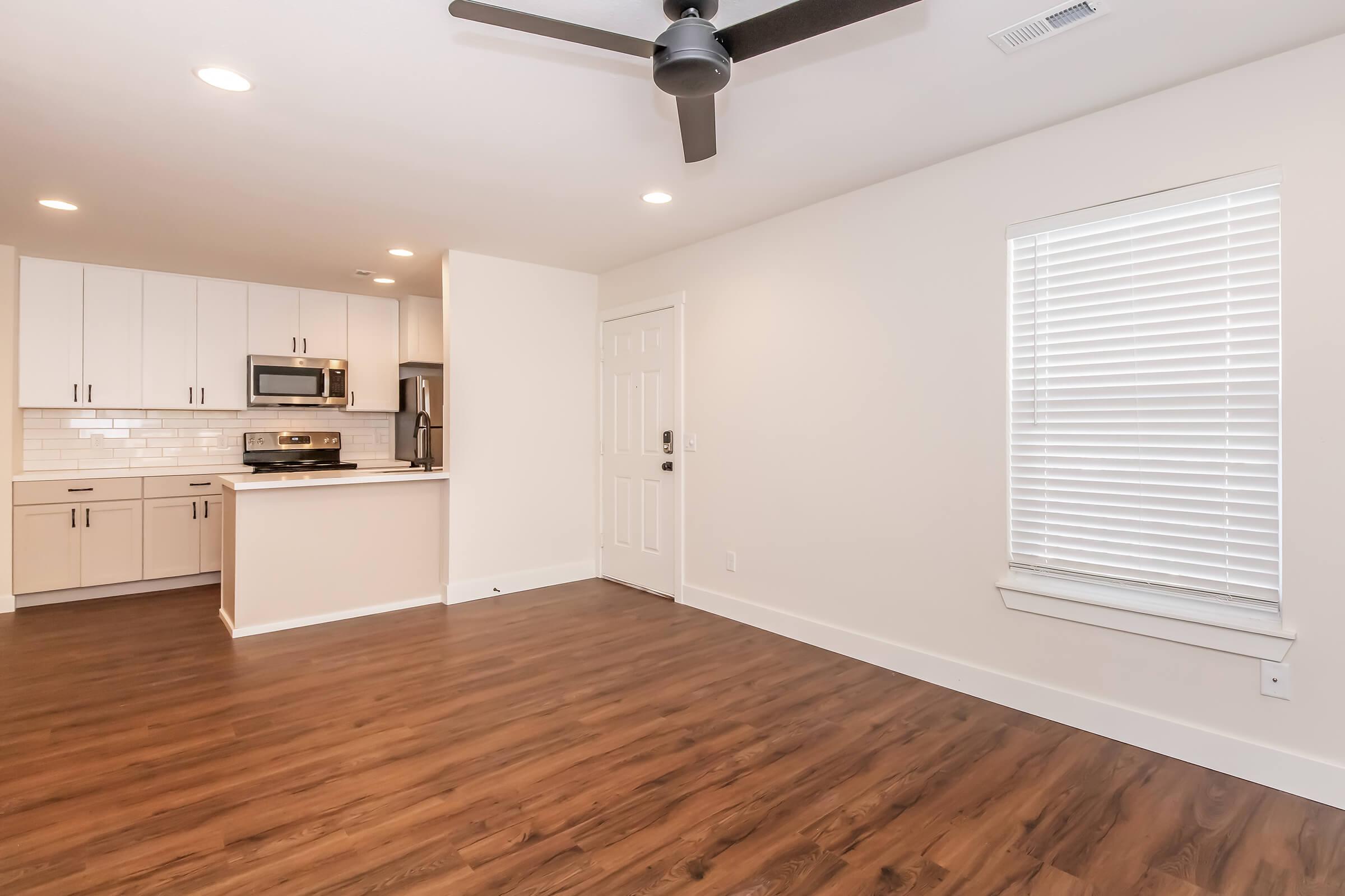 Interior view of a modern apartment living room featuring hardwood flooring, a ceiling fan, and a large window with blinds. An adjacent kitchen area includes white cabinets, stainless steel appliances, and a tile backsplash. The space is well-lit and designed in a contemporary style.