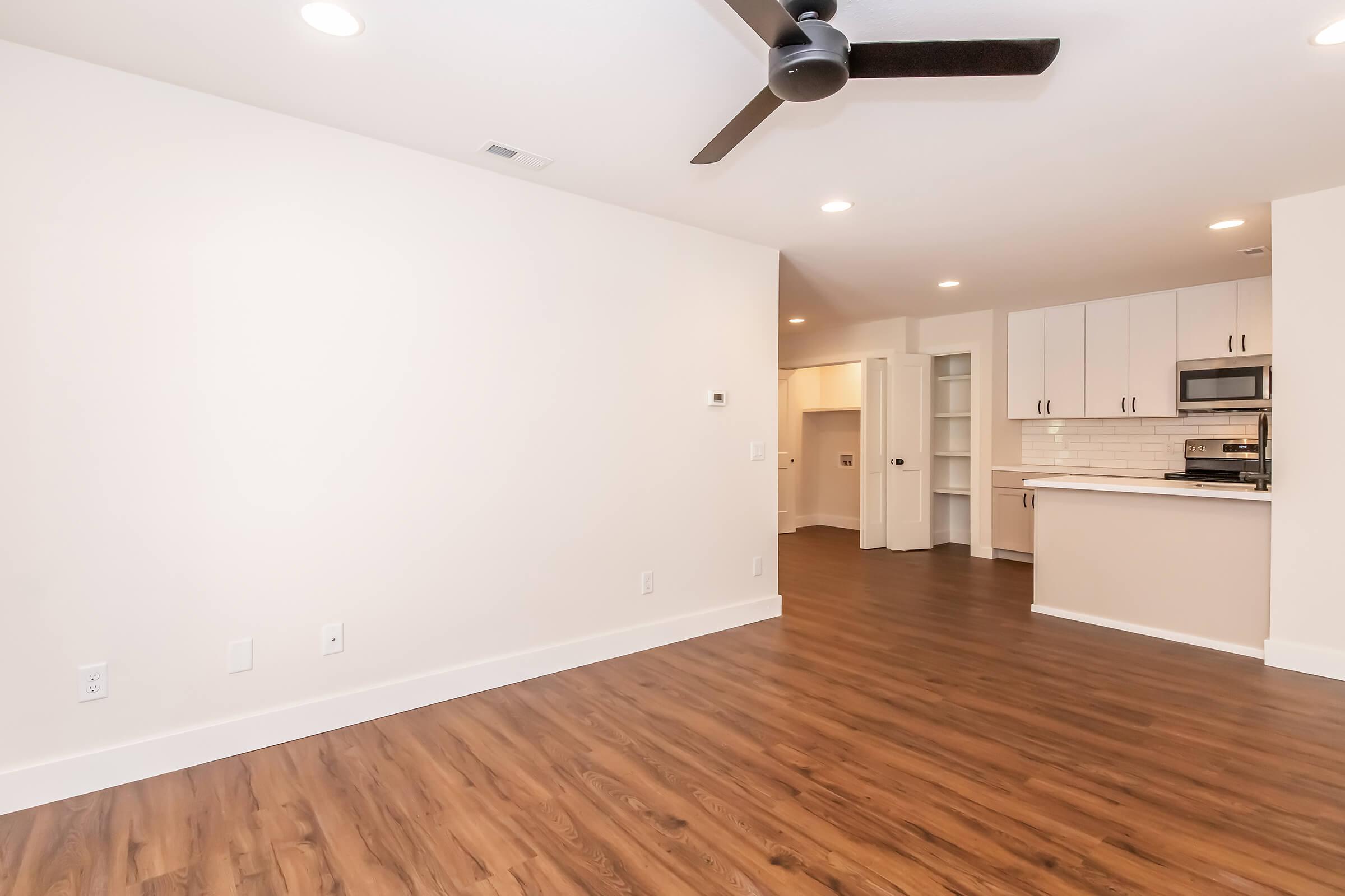 A modern, light-filled room with a ceiling fan and wooden flooring. The space features neutral-colored walls and a kitchenette area with a sleek design, including a stainless steel microwave. There are built-in shelves in the background, enhancing the overall contemporary look.