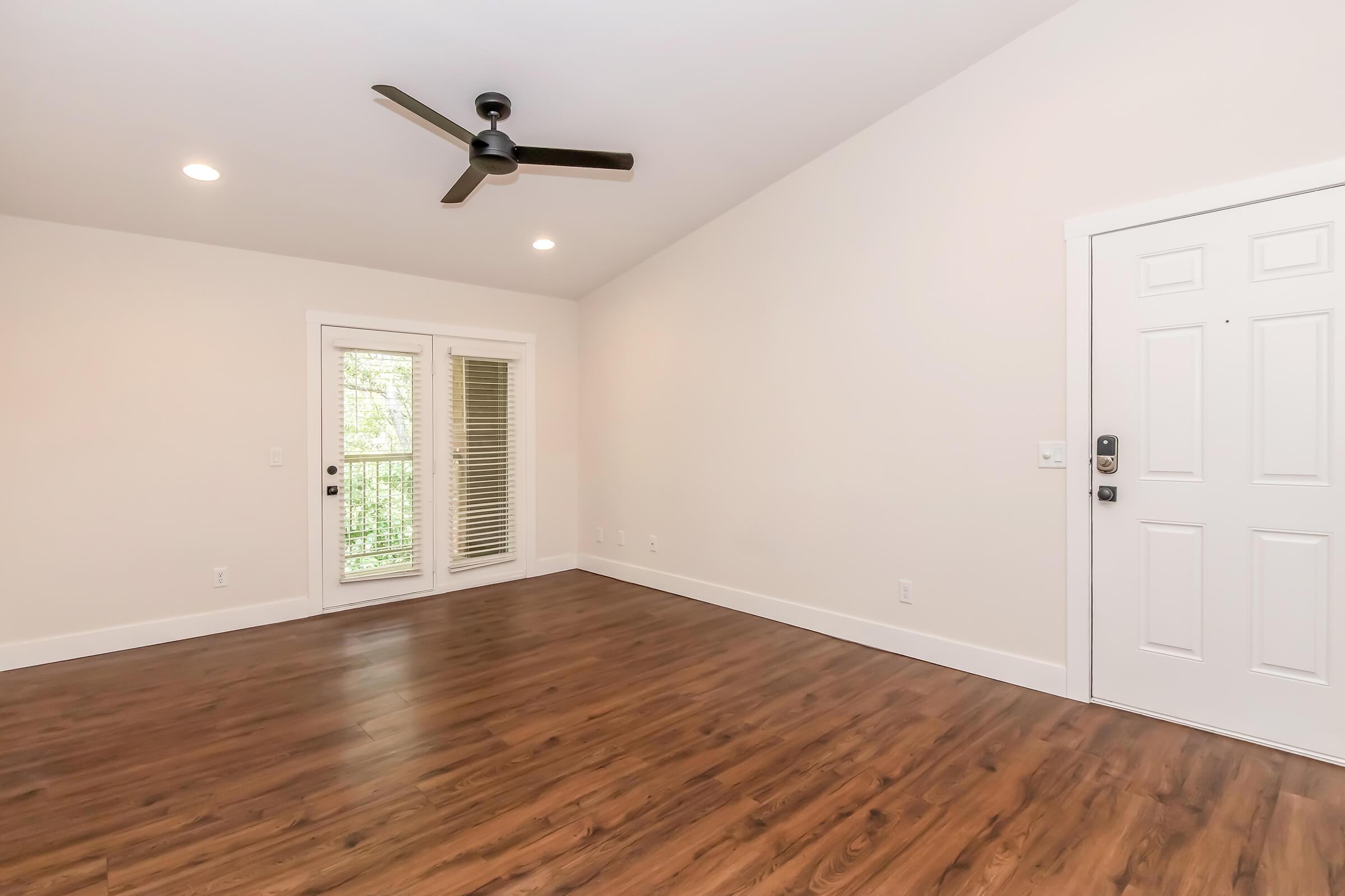 A spacious, brightly lit living room featuring hardwood floors, a ceiling fan, a pair of sliding glass doors leading to a balcony, and a white front door. The walls are painted in a soft beige color, creating a warm and inviting atmosphere.