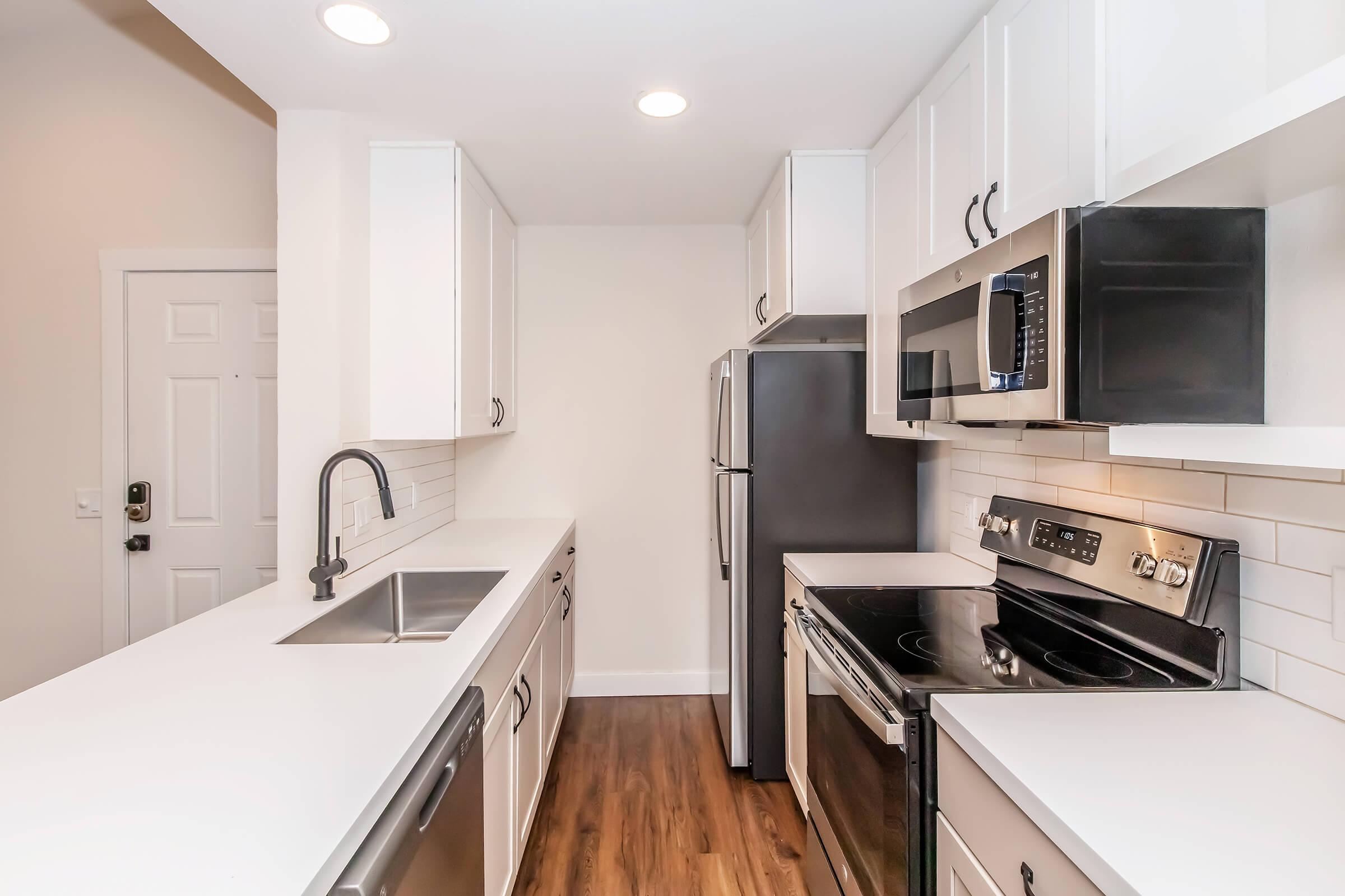 Modern kitchen featuring white cabinets, stainless steel appliances including a microwave, oven, and refrigerator, along with a sleek countertop and a sink. The layout is open and well-lit with recessed lighting, and it has a clean, minimalist design with wooden flooring.