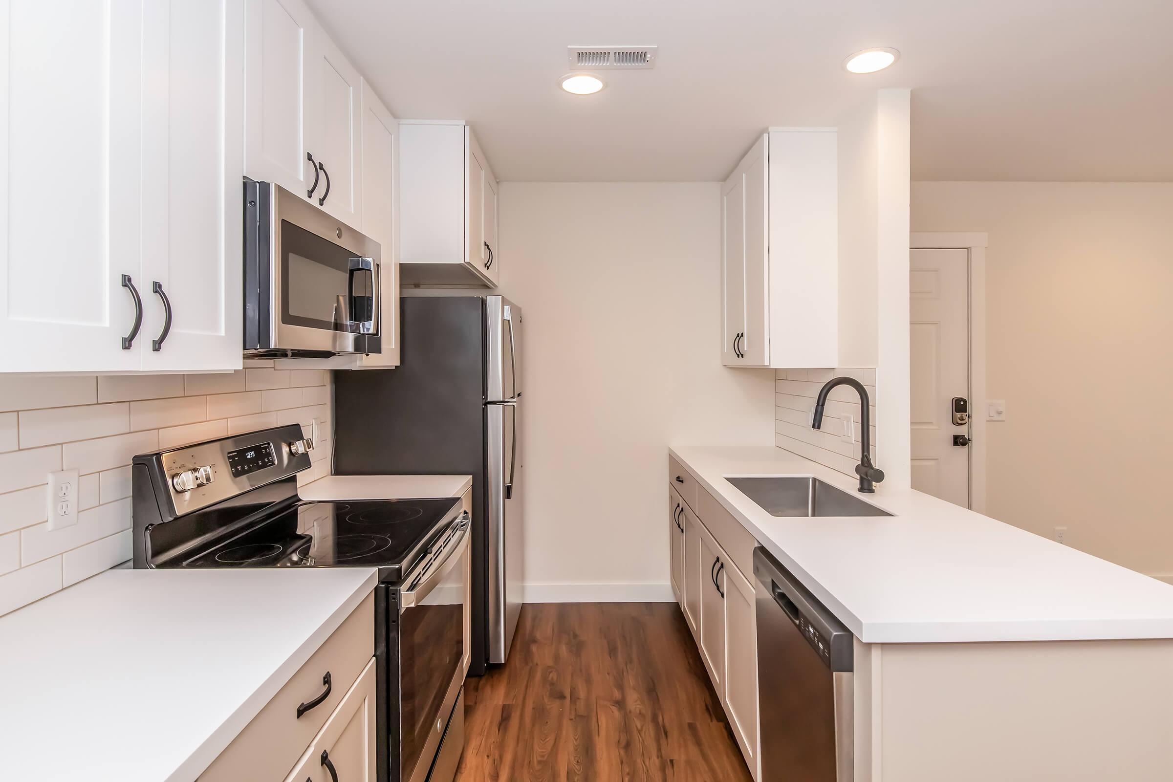 Modern kitchen featuring white cabinets, stainless steel appliances including a microwave, refrigerator, and oven, and a sleek countertop. The kitchen has a contemporary design with a large sink, ample counter space, and warm wood flooring, creating a clean and inviting atmosphere.