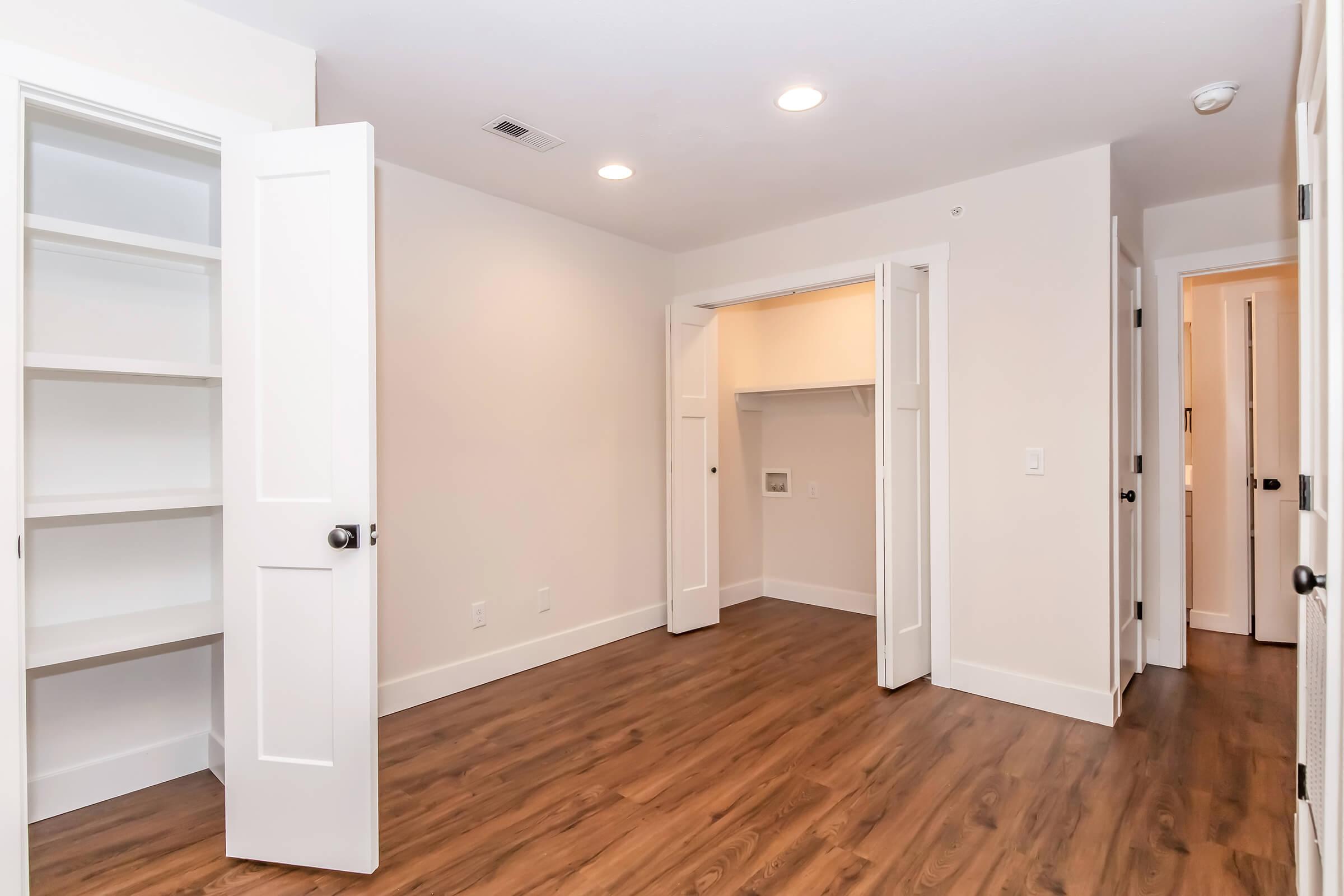 A spacious, empty room featuring light-colored walls, hardwood flooring, and multiple doors. One door leads to a closet space, while another reveals a small nook with shelves. The room has overhead lighting and a neutral decor, creating a bright and airy atmosphere.
