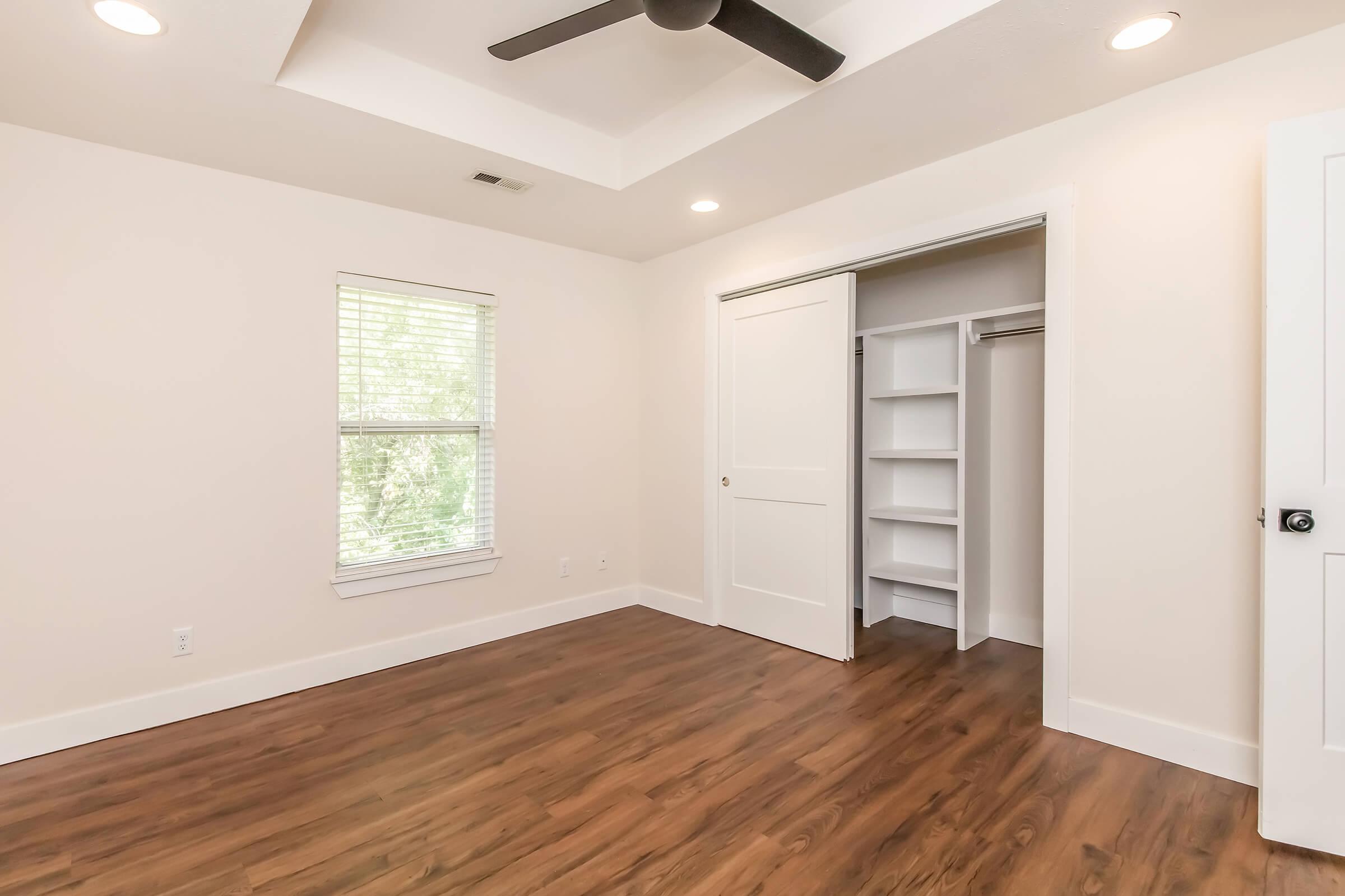 A well-lit empty room featuring light-colored walls, a ceiling fan, and a window with blinds. There is a closet with sliding doors on the right, and the floor is covered in warm-toned wooden laminate. The overall ambiance is clean and modern.