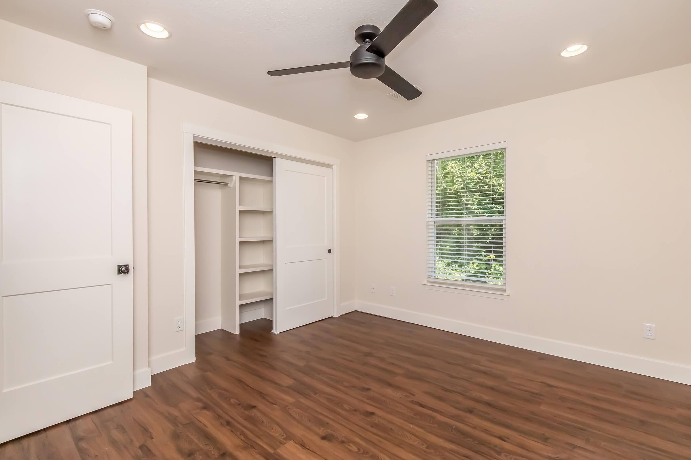 A spacious, empty room with light-colored walls, a ceiling fan, and brown wood-like flooring. It features a window with blinds allowing natural light, a closet with sliding doors, and white trim around the doors and window. The overall atmosphere is bright and airy, suitable for various uses.