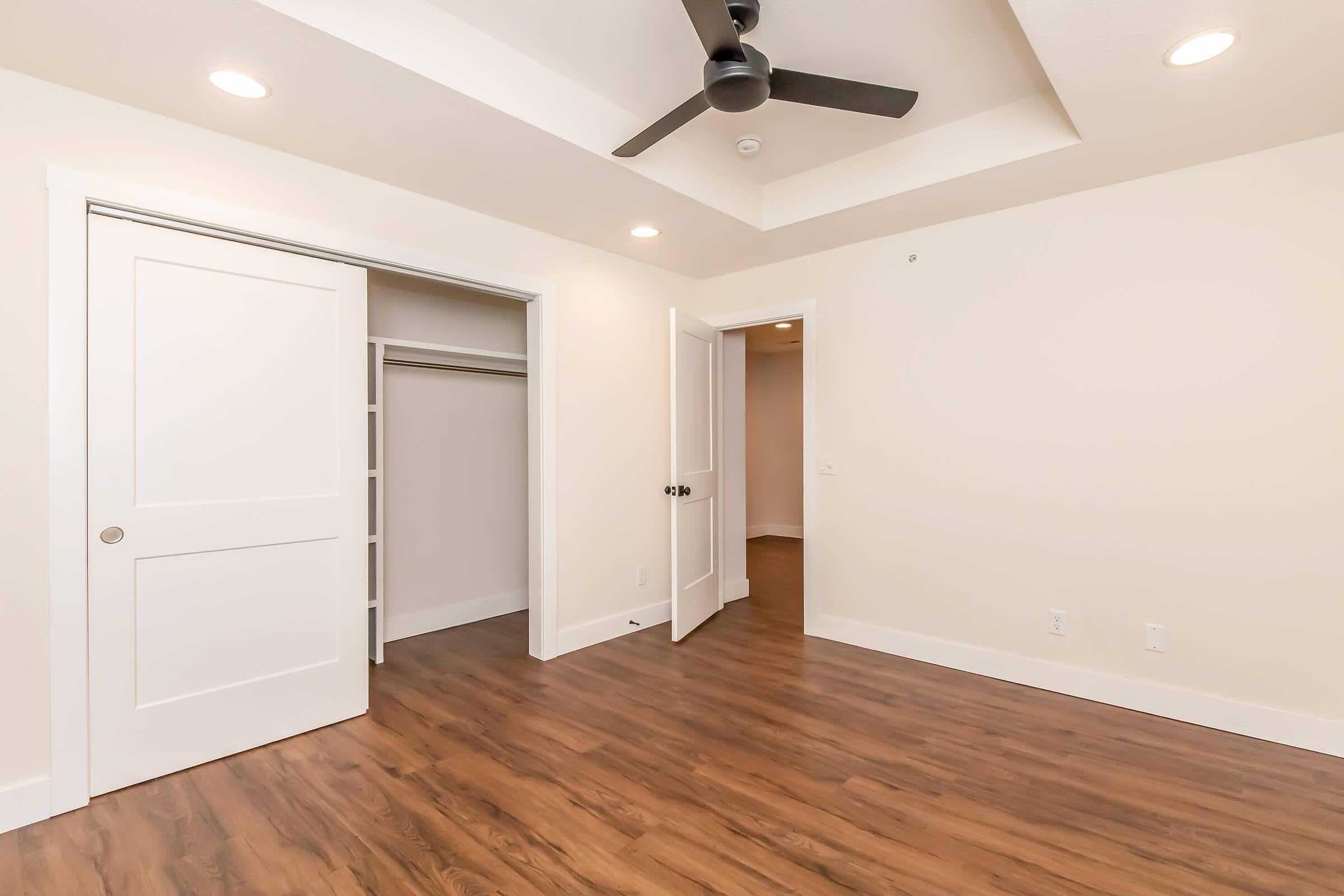 A modern, empty room with beige walls and dark hardwood flooring. There are two white doors; one leading to a closet and the other to another space. A ceiling fan is installed above, and the room is brightly lit, creating a spacious feel.
