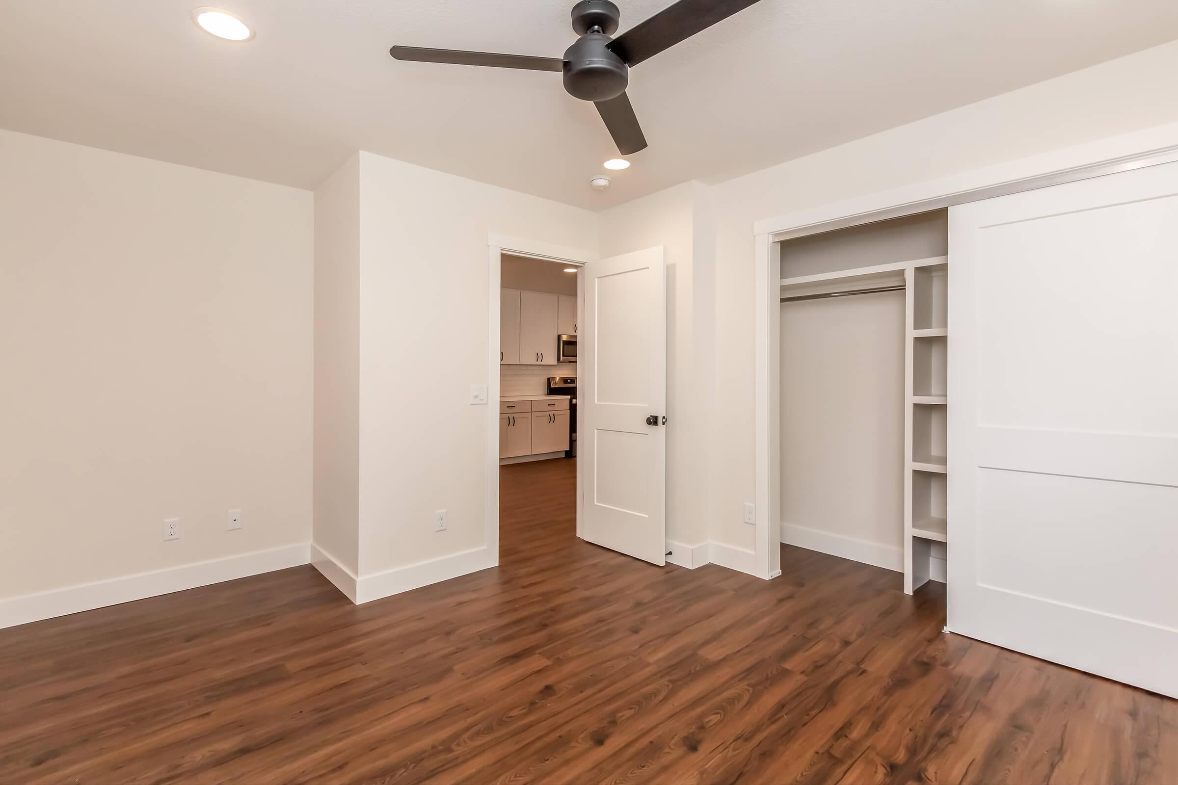 A well-lit room with light walls and laminate wood flooring. It features a ceiling fan, a closet with sliding doors, and an open doorway leading to another area. The space is neat and modern, suitable for a bedroom or office.
