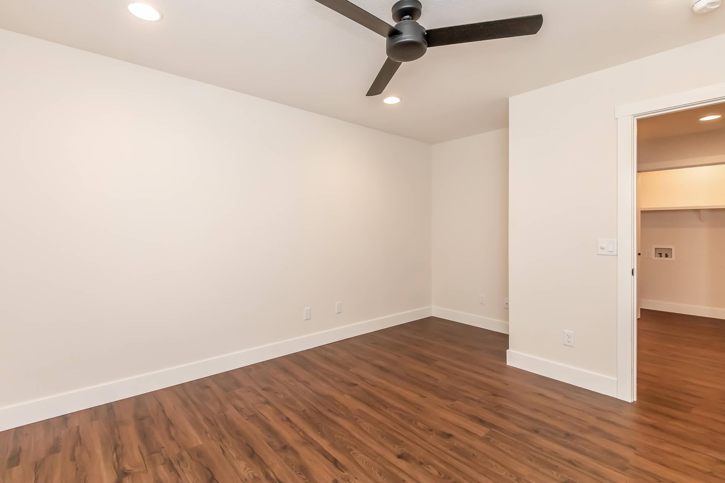 An empty room featuring light-colored walls, a ceiling fan, and hardwood flooring. There is a doorway on the right leading to a closet or another space, and ceiling lights provide illumination. The room is spacious and has a minimalist design.
