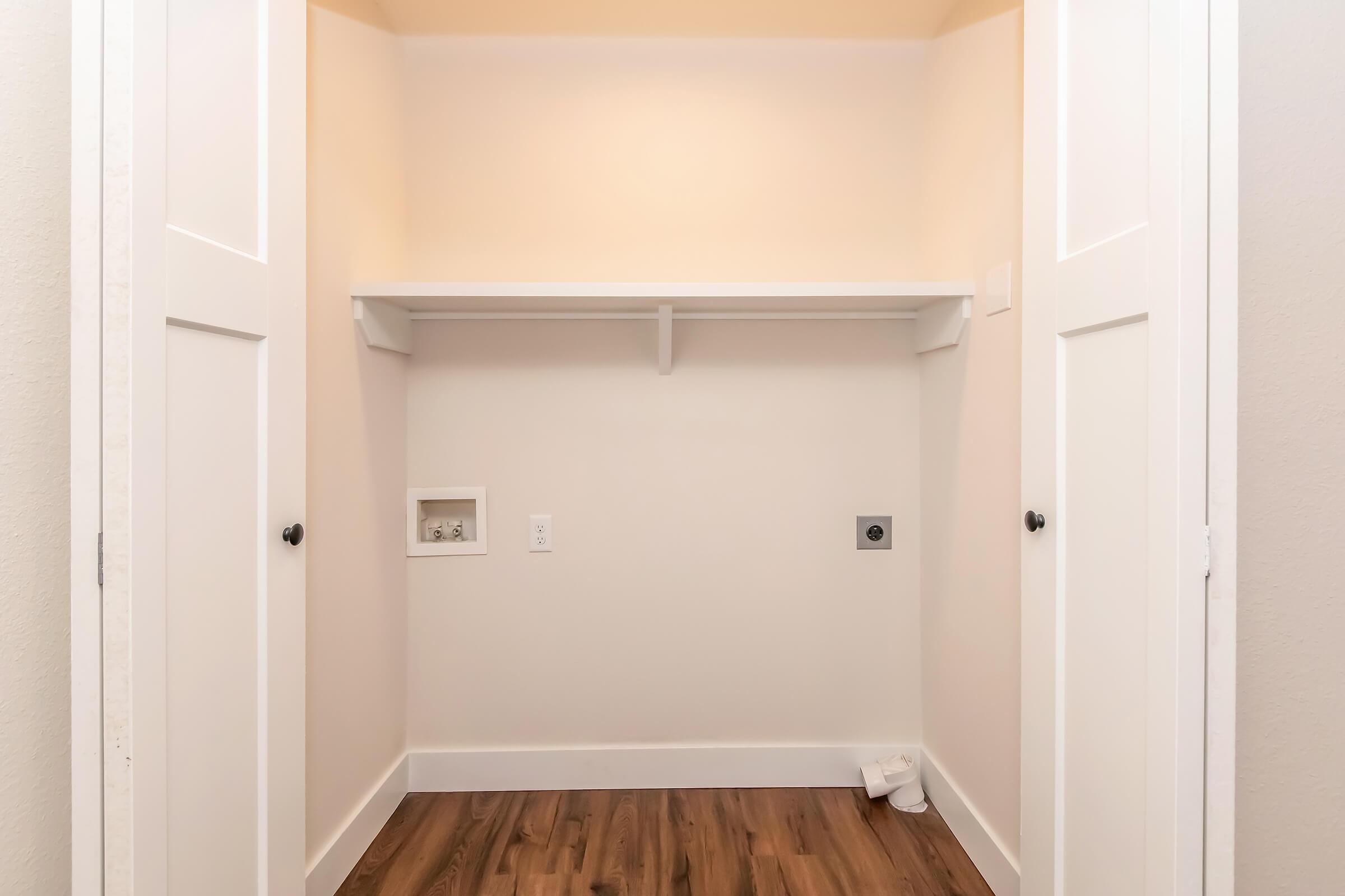 A vacant closet with light-colored walls and a wooden floor. It features double doors, a single shelf along the top, and a small wall outlet. There’s one white item on the floor, but the space is mostly empty, suggesting it is ready for storage or organization.