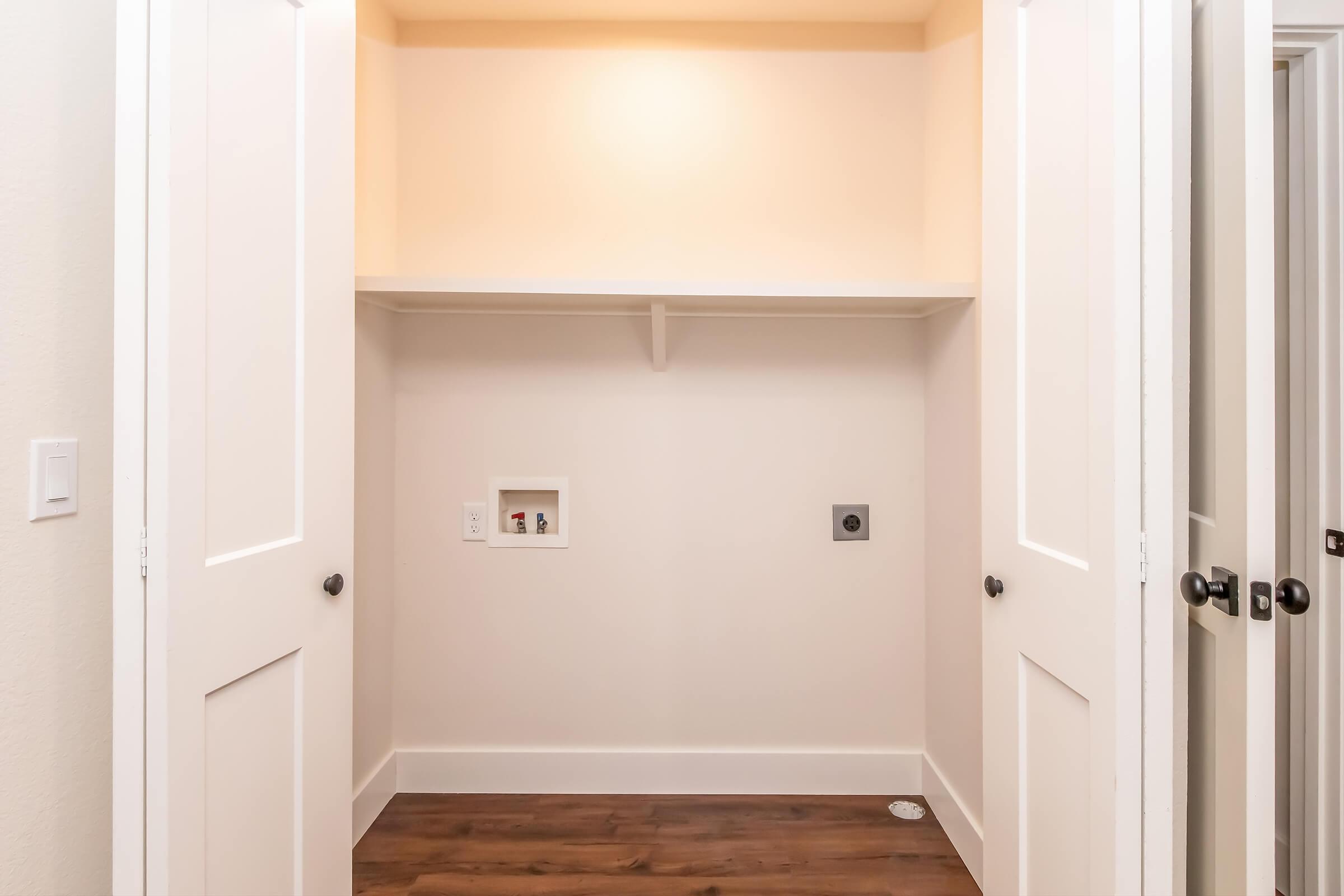 A clean, empty closet with two white doors partially open. It features a single shelf above a small recess with electrical outlets, and a dark wood floor. The walls are painted a light color, creating a spacious and minimalistic appearance.