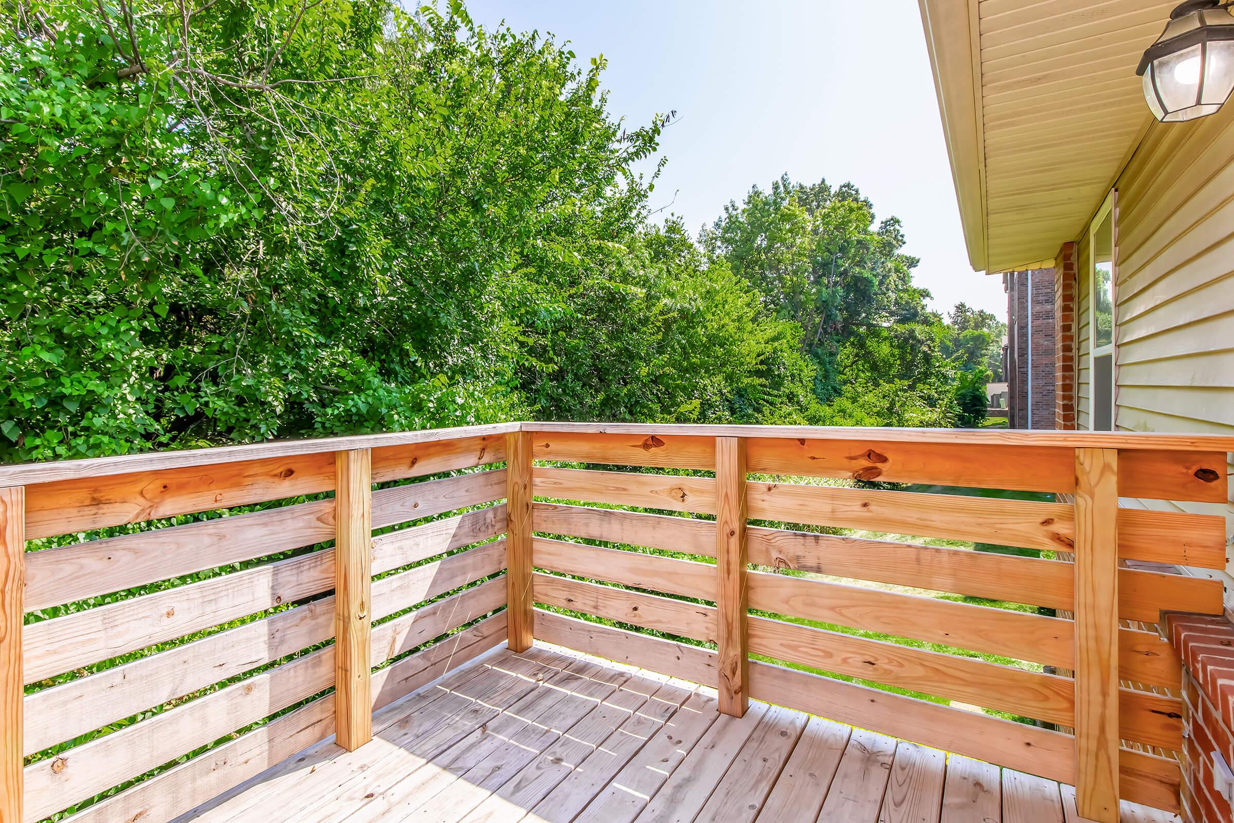 A wooden balcony with a railing, overlooking a lush green area with trees. The floor is made of wooden planks, and the balcony is attached to a house with a light fixture on the wall. Bright sunlight creates a warm atmosphere, highlighting the natural surroundings.