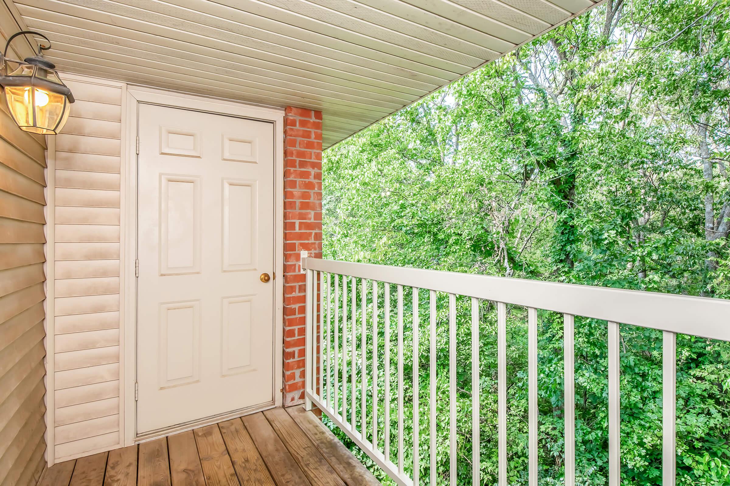A balcony view featuring a wooden floor and a white railing, with a closed white door on the left and a brick wall. Lush green trees and foliage are visible in the background, creating a natural and serene atmosphere. A lantern hangs near the door, adding to the charm of the space.