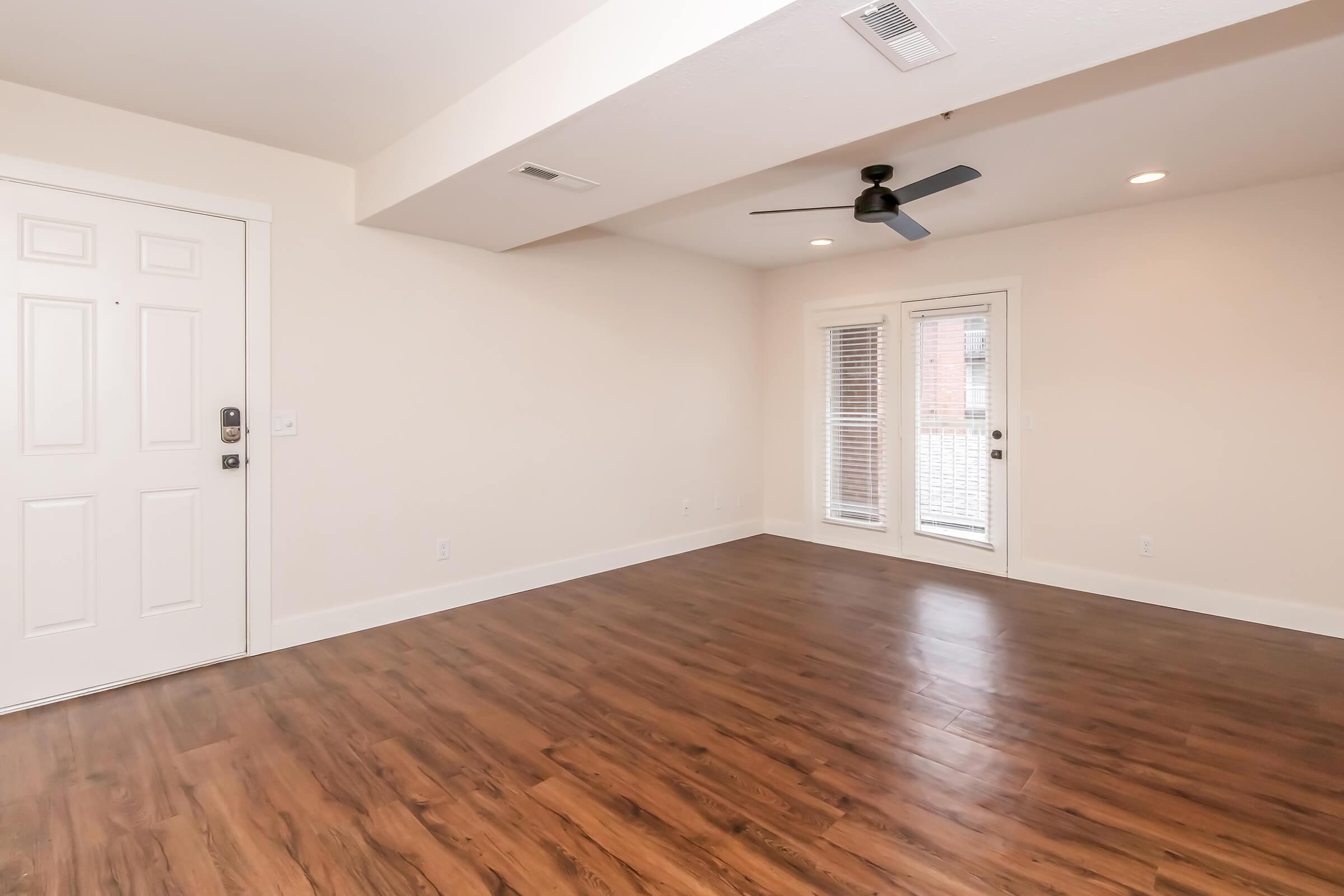 Spacious interior of a bright room featuring light-colored walls, a ceiling fan, and wood-style flooring. Includes a front door and glass doors leading to an outdoor space, allowing natural light to fill the area.