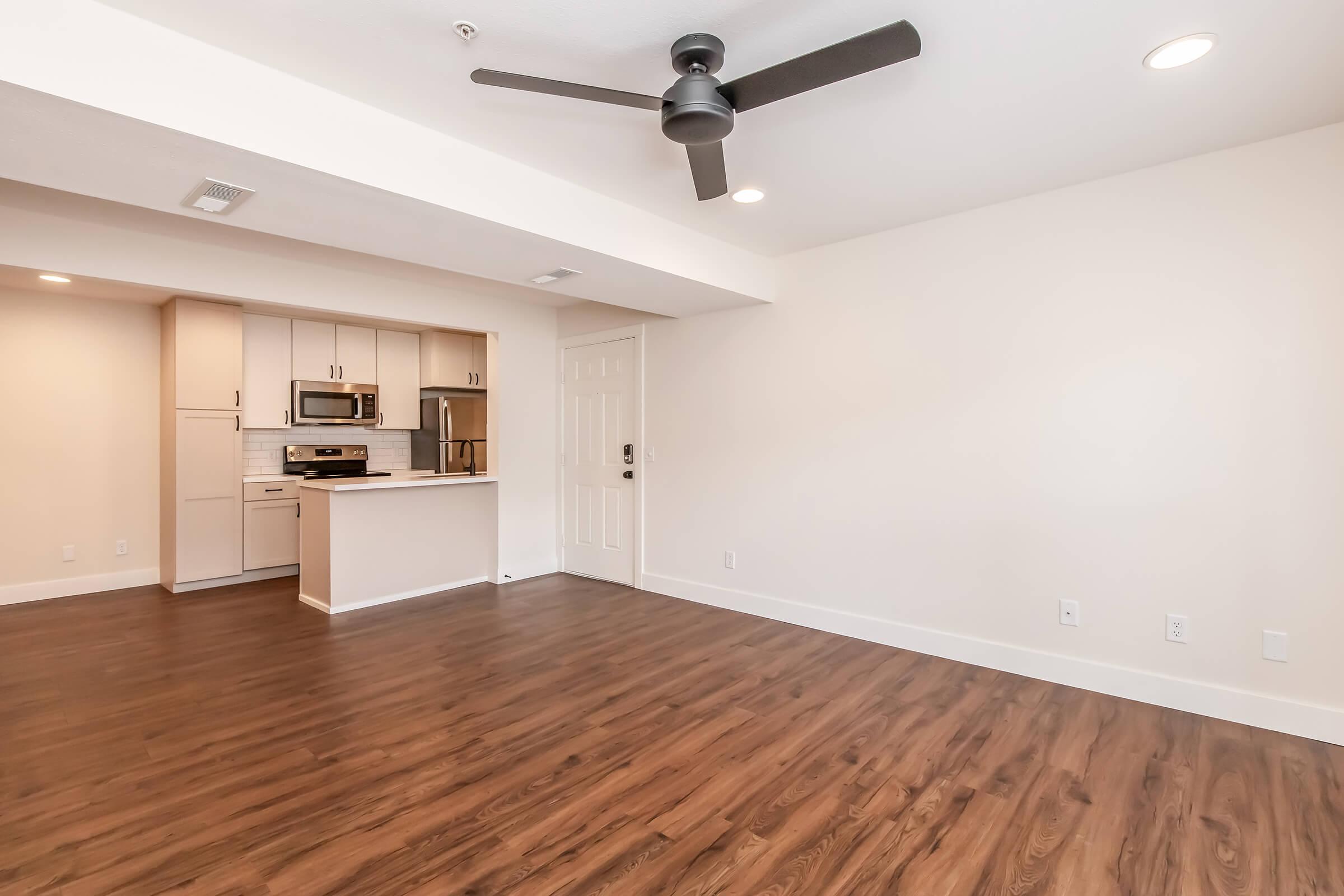 Spacious modern living area featuring hardwood floors, a ceiling fan, and an open kitchen with stainless steel appliances and a breakfast bar. The walls are painted in a light color, providing a bright and airy atmosphere.