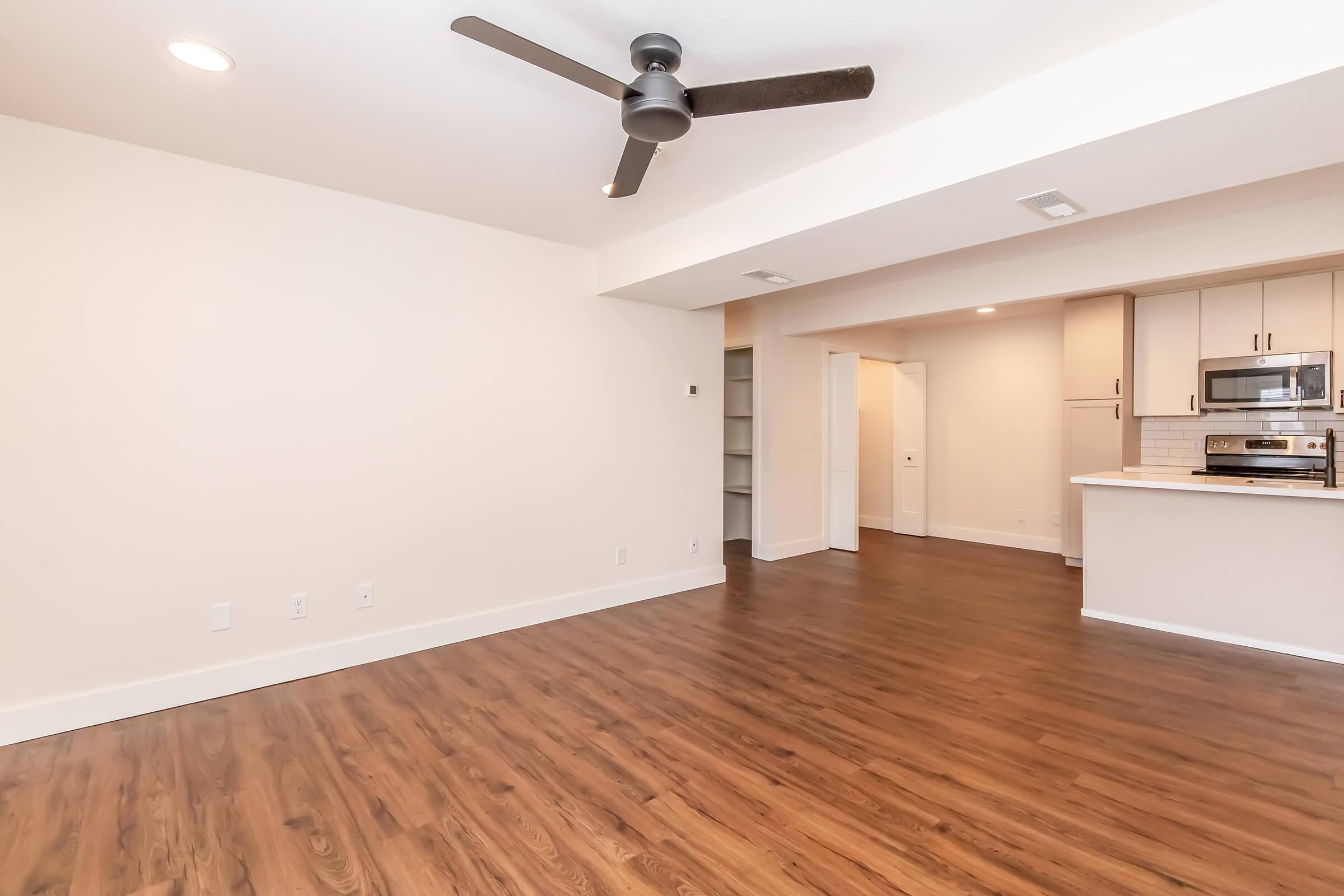 A spacious, well-lit empty room with a light wooden floor and a ceiling fan. The walls are painted in a neutral color. To the right, there is a modern kitchen area featuring stainless steel appliances and an open layout. There are doorways leading to other areas, suggesting a functional living space.