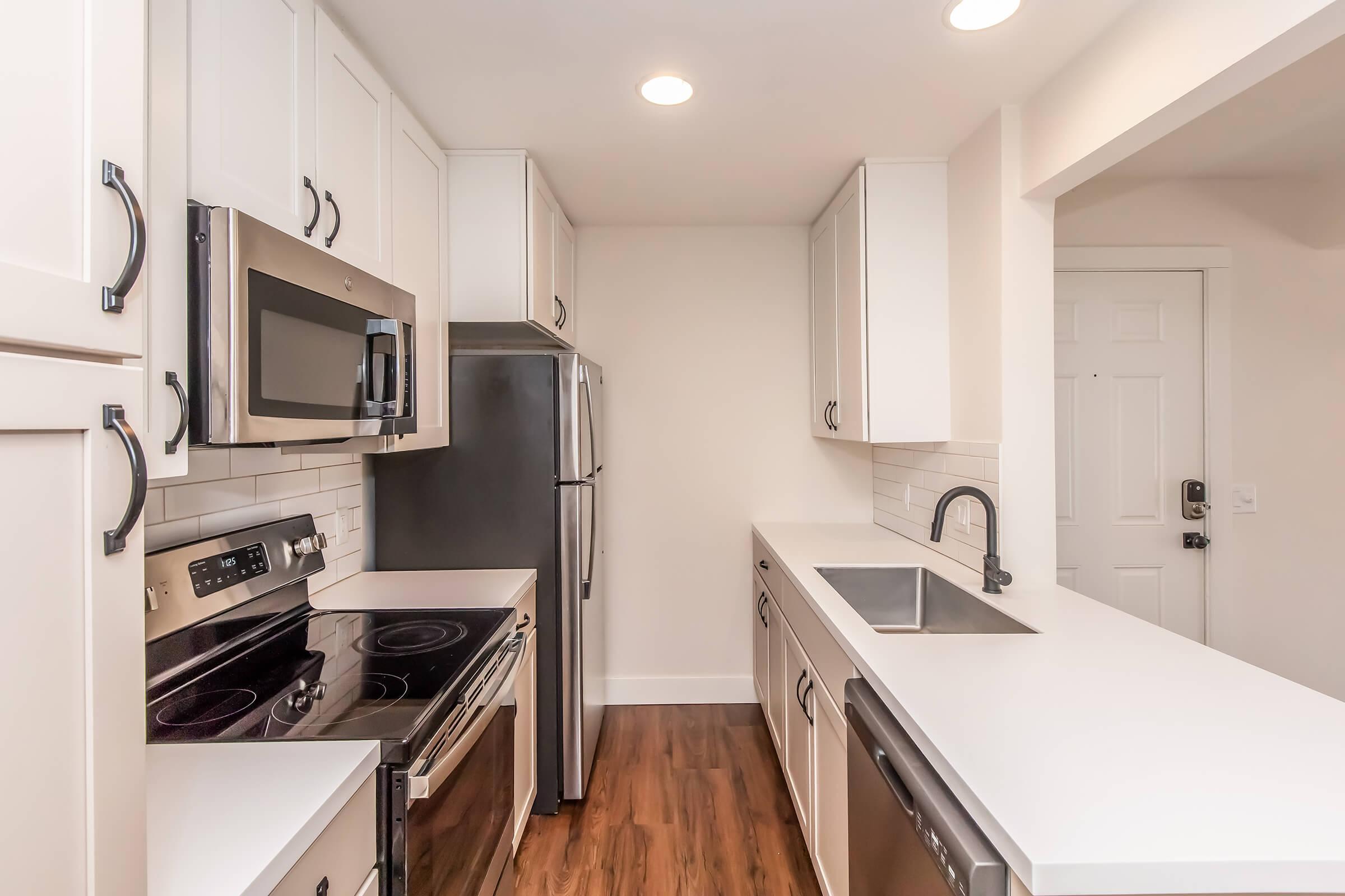 Modern kitchen featuring white cabinets, stainless steel appliances including a microwave, refrigerator, and oven. The countertop is sleek and white, with a sink and a black faucet. The flooring is a warm wood laminate. Soft overhead lighting illuminates the space with an open layout leading to an entrance door.