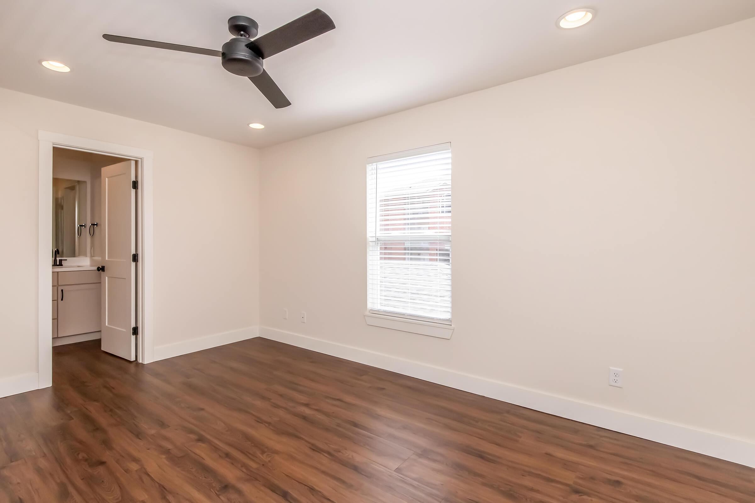 Spacious room with light-colored walls and brown wooden flooring. A ceiling fan is mounted overhead. There is a window with blinds allowing natural light, and a doorway leading to an adjacent bathroom. The overall ambiance is modern and inviting.