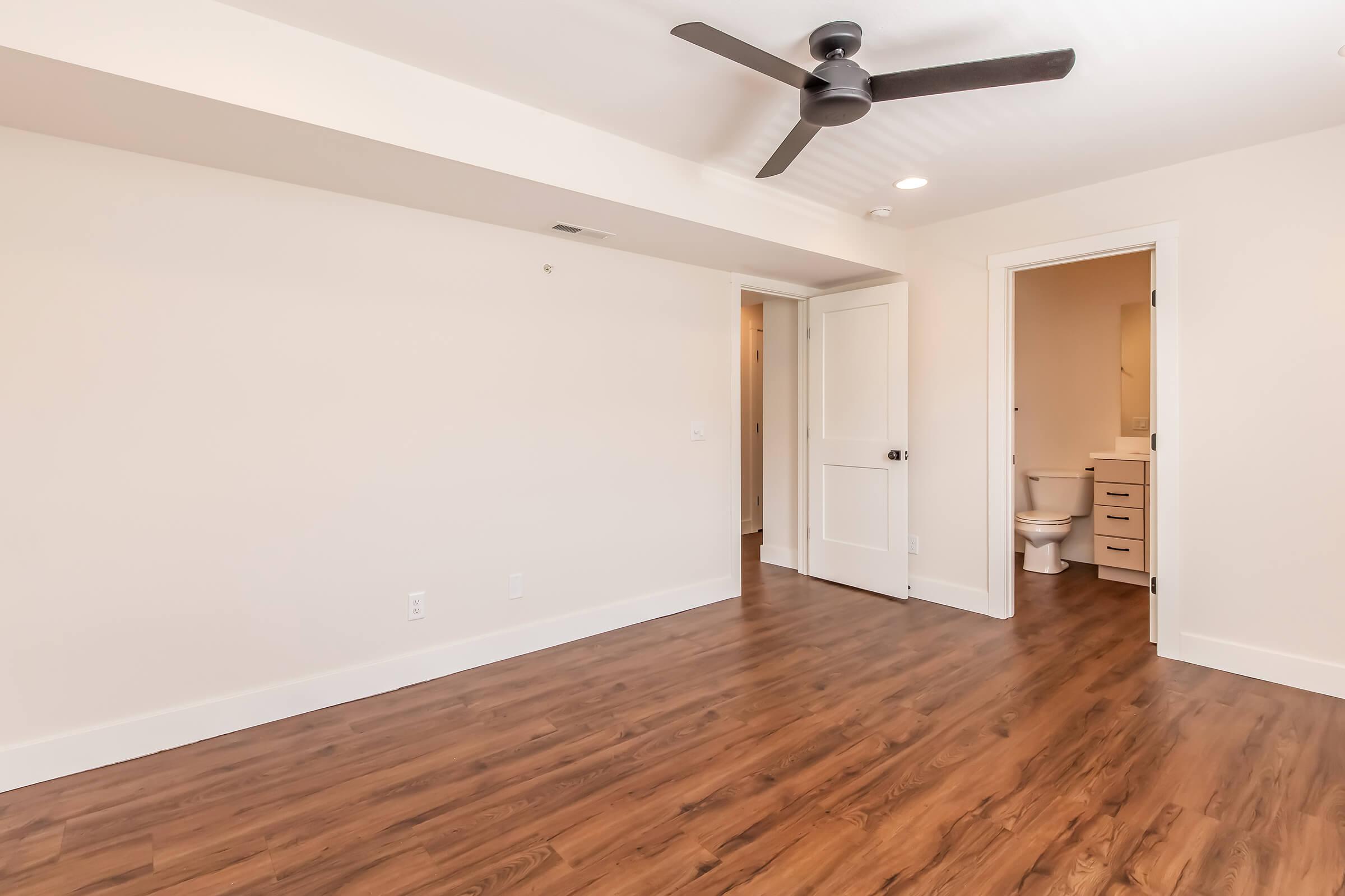 A well-lit interior space featuring light-colored walls and a wooden floor. A ceiling fan is overhead. In the background, there is a doorway leading to a bathroom with a toilet. The room appears empty, providing a clean and modern aesthetic.