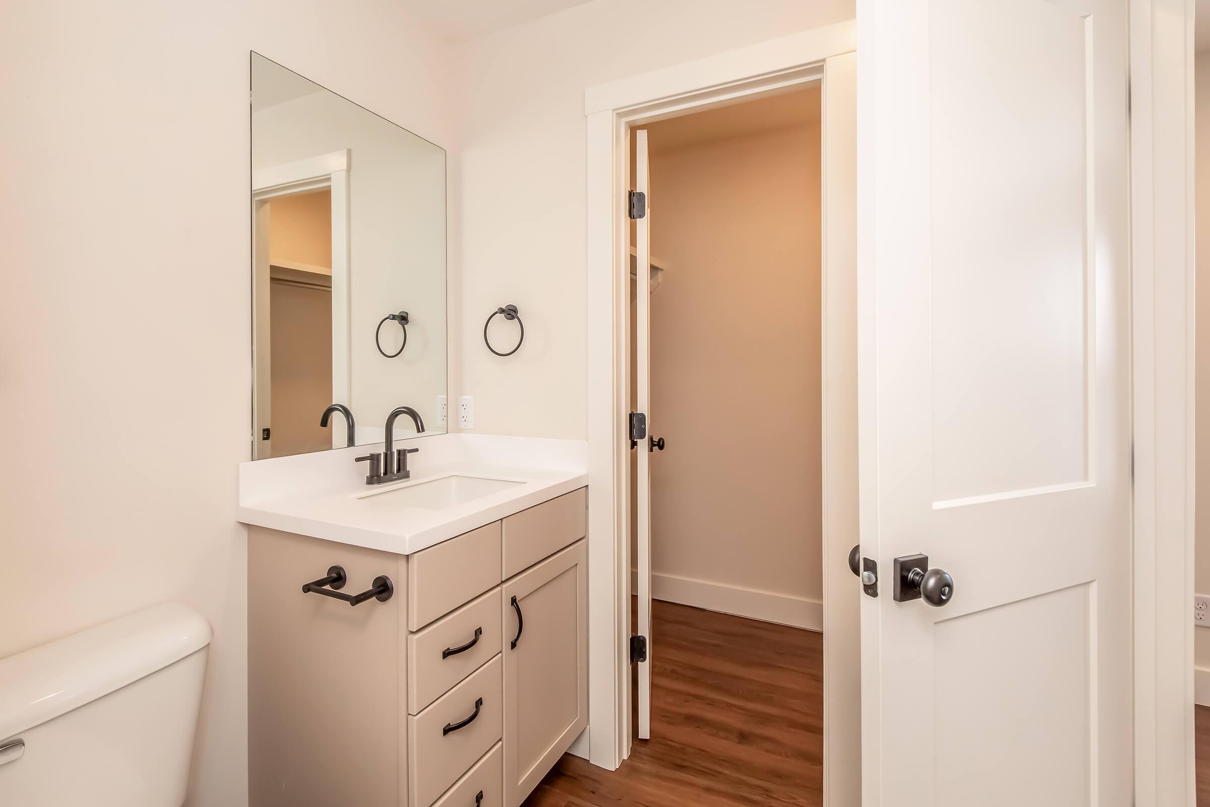 A modern bathroom featuring a stylish vanity with a white countertop and black faucet, a large mirror above it, and towel rings. To the right, there's an open door leading to another room, with light-colored walls and wooden flooring throughout.