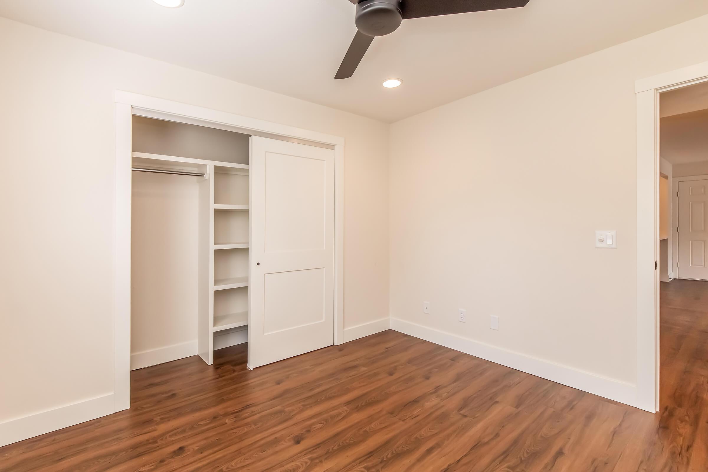 A well-lit, minimalist room featuring a hardwood floor, a ceiling fan, and white walls. There is an open closet with built-in shelves to the left, and a closed door on the right leading to another space. The overall atmosphere is clean and modern.