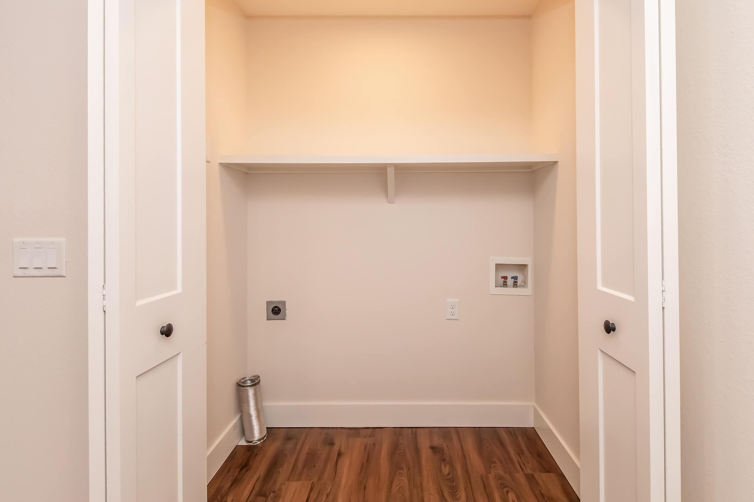 Interior of an unoccupied closet featuring two white doors, a shelf along the back wall, and finished wood flooring. There are electrical outlets and a vent on the wall, along with a metallic cylinder on the floor. The walls and ceiling are painted a soft light color.