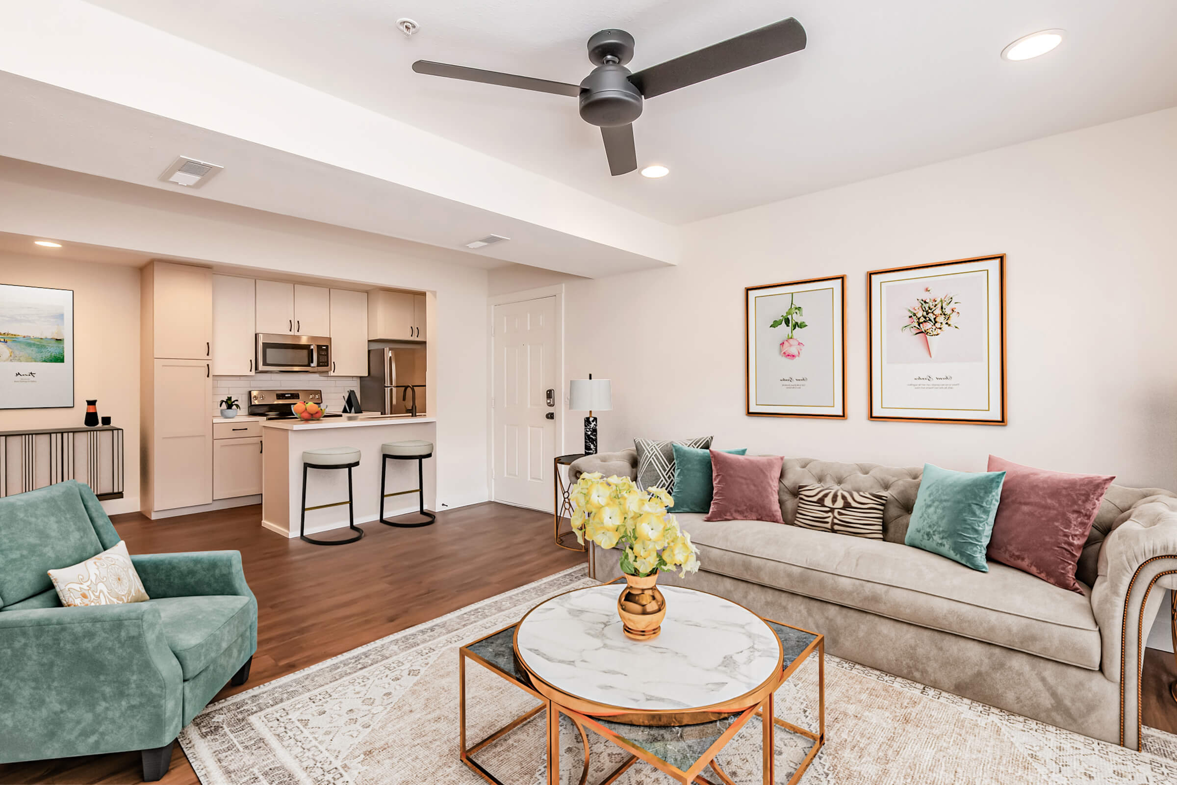 A modern living room featuring a light-colored sofa with decorative pillows, a green accent chair, and a round coffee table with a floral centerpiece. The space includes framed botanical artwork on the walls and a kitchen visible in the background, creating a stylish and inviting atmosphere.