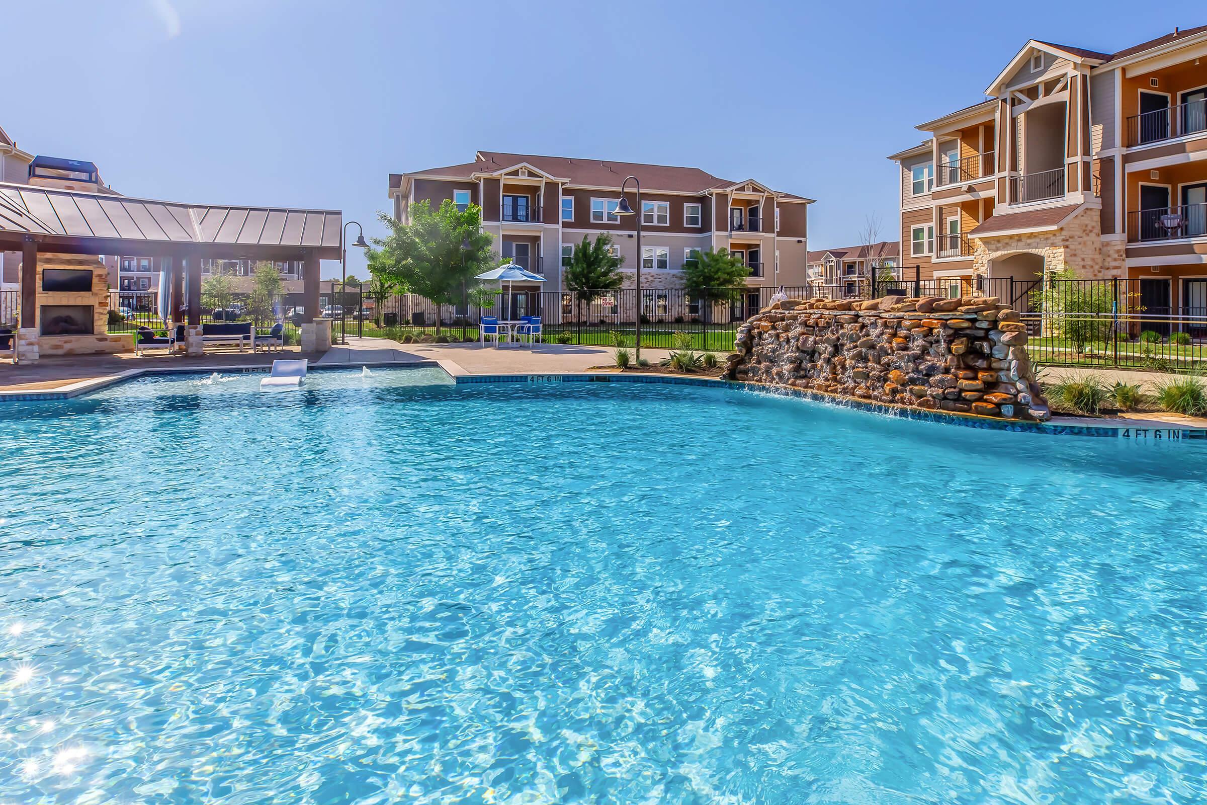 A serene swimming pool surrounded by landscaped greenery and residential buildings. The pool features a diving board and a stone waterfall, creating a relaxing atmosphere under clear blue skies. Sunny conditions highlight the shimmering water, inviting residents to enjoy outdoor leisure.