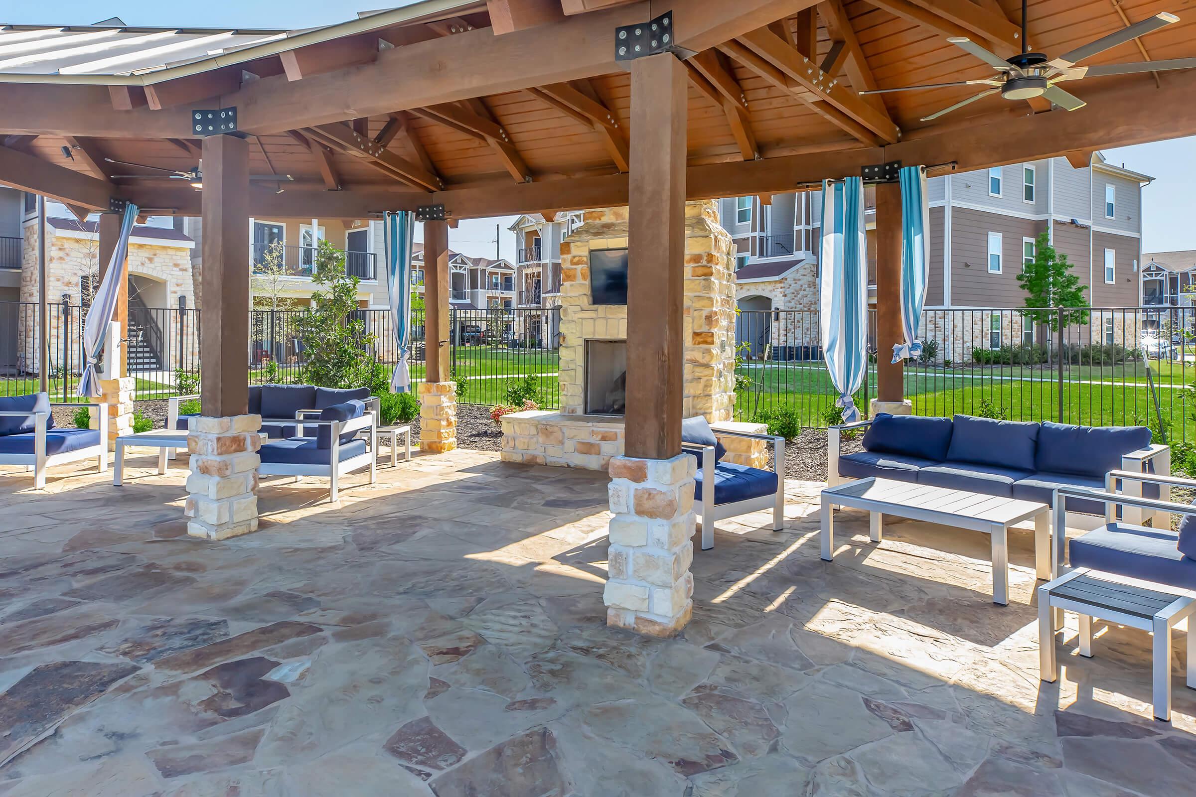A shaded outdoor seating area with wooden beams and stone accents, featuring cushioned sofas and a coffee table. In the background, there are other buildings and greenery. A cozy, inviting space for relaxation or social gatherings, with a fireplace and ceiling fans for comfort.